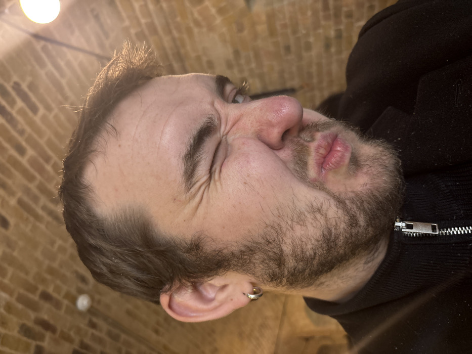A close-up, slightly tilted shot of a man with a beard and dark hair, wearing a black zip-up jacket. His face is scrunched up, suggesting a squint or grimace. The background appears to be a brick wall. A bright light source is visible in the upper left corner. The man has a silver earring in his left ear. His expression is the most prominent aspect of the image.