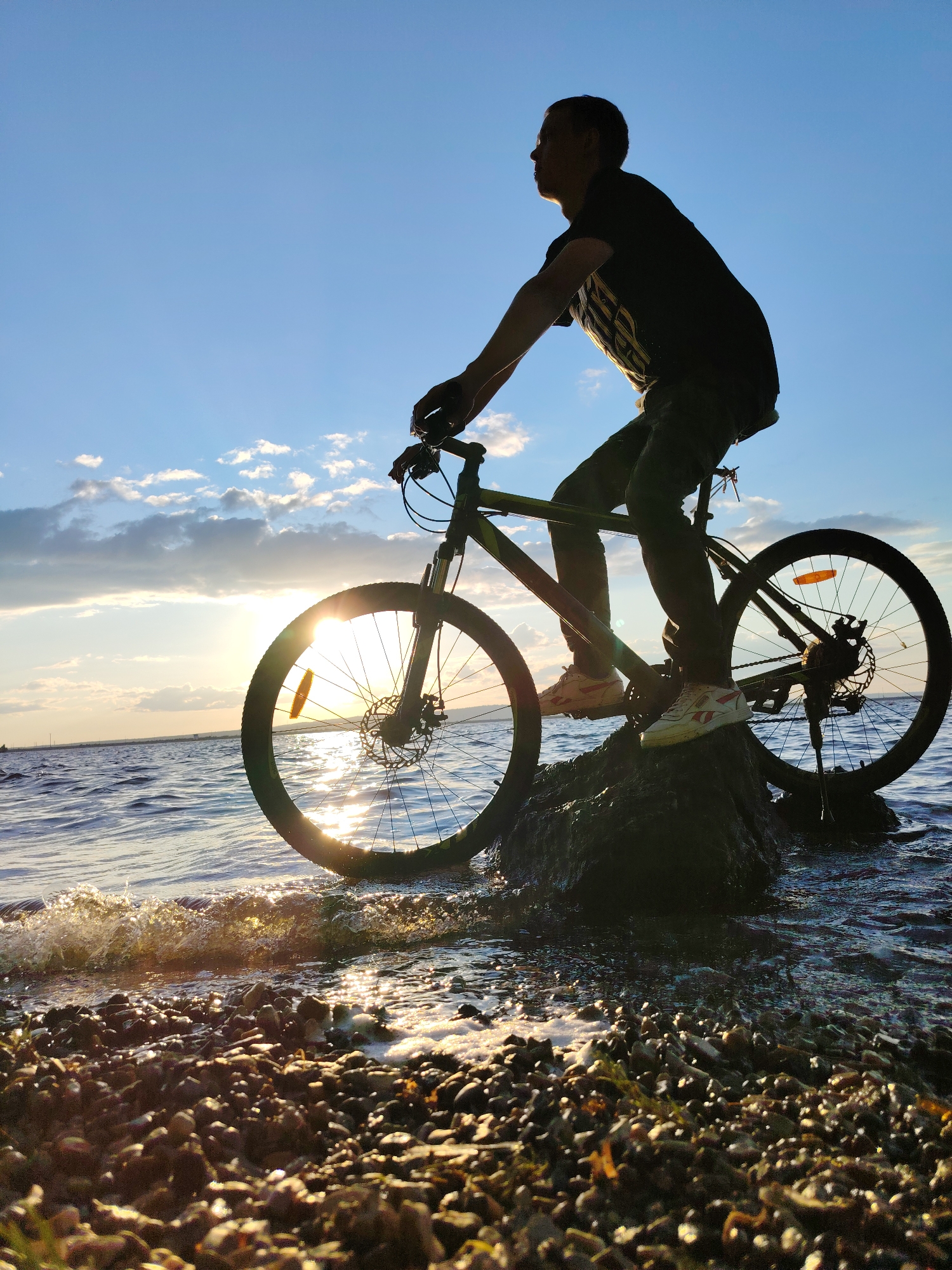 A silhouette of a person on a bicycle stands on a rock partially submerged in water, with the sun setting in the background. The person is wearing a dark t-shirt and pants, and the bicycle appears to be a mountain bike. The water is calm with small waves lapping at the shore, which is covered in pebbles. The sky is blue with scattered clouds, and the sun is low on the horizon, casting a warm glow. The location is the Kama River embankment in Chaikovsky, Russia.
