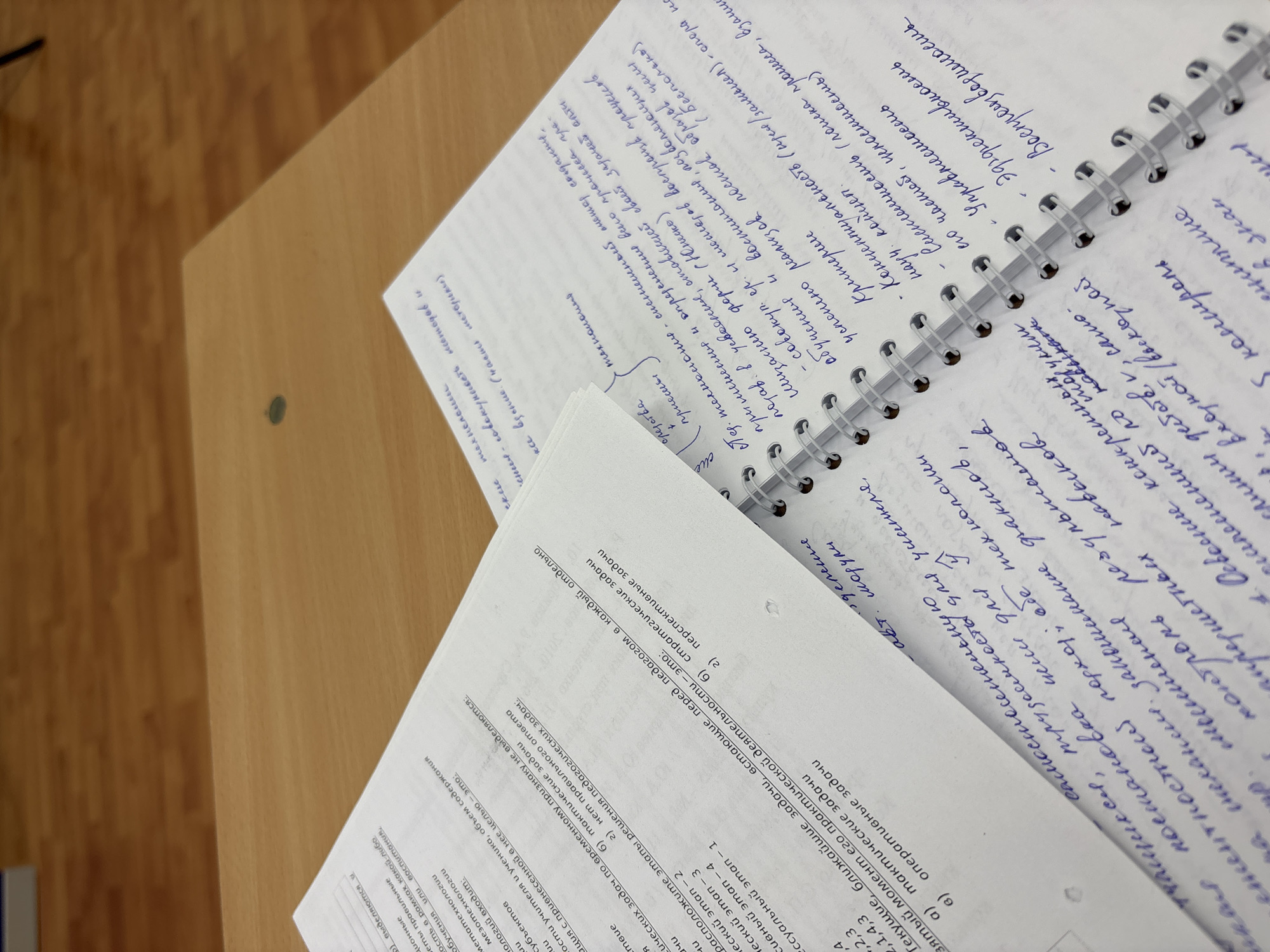 The image captures a close-up view of study materials on a desk, likely in an educational setting. The focus is on two documents: a handwritten page from a spiral-bound notebook and a printed handout. The handwritten notes are in Russian, densely filled with text and annotations in blue ink. The printed handout, also in Russian, features a list of numbered questions or points, possibly related to pedagogy or educational technology. The desk surface has a wood-like finish, and the overall lighting suggests an indoor environment. The items are closely arranged, giving a sense of active study or preparation. The location is implied by the language of the text to be in Russia, possibly in the city of Chaikovsky.