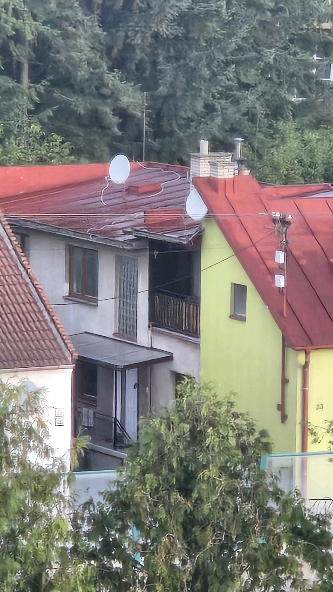 The image shows a residential building with a red roof, situated amidst lush green trees. The building features a prominent yellow facade on the right side, and a lighter colored facade on the left. Several satellite dishes are visible on the roof. A chimney stack is present on the roof of the yellow section. A balcony with wooden railings is visible on the left side of the yellow section. The building appears to be located in a residential area of Prešov, Slovakia. The time of day appears to be daytime, with the lighting suggesting it could be late afternoon. The overall impression is that of a quiet, leafy suburban neighborhood.