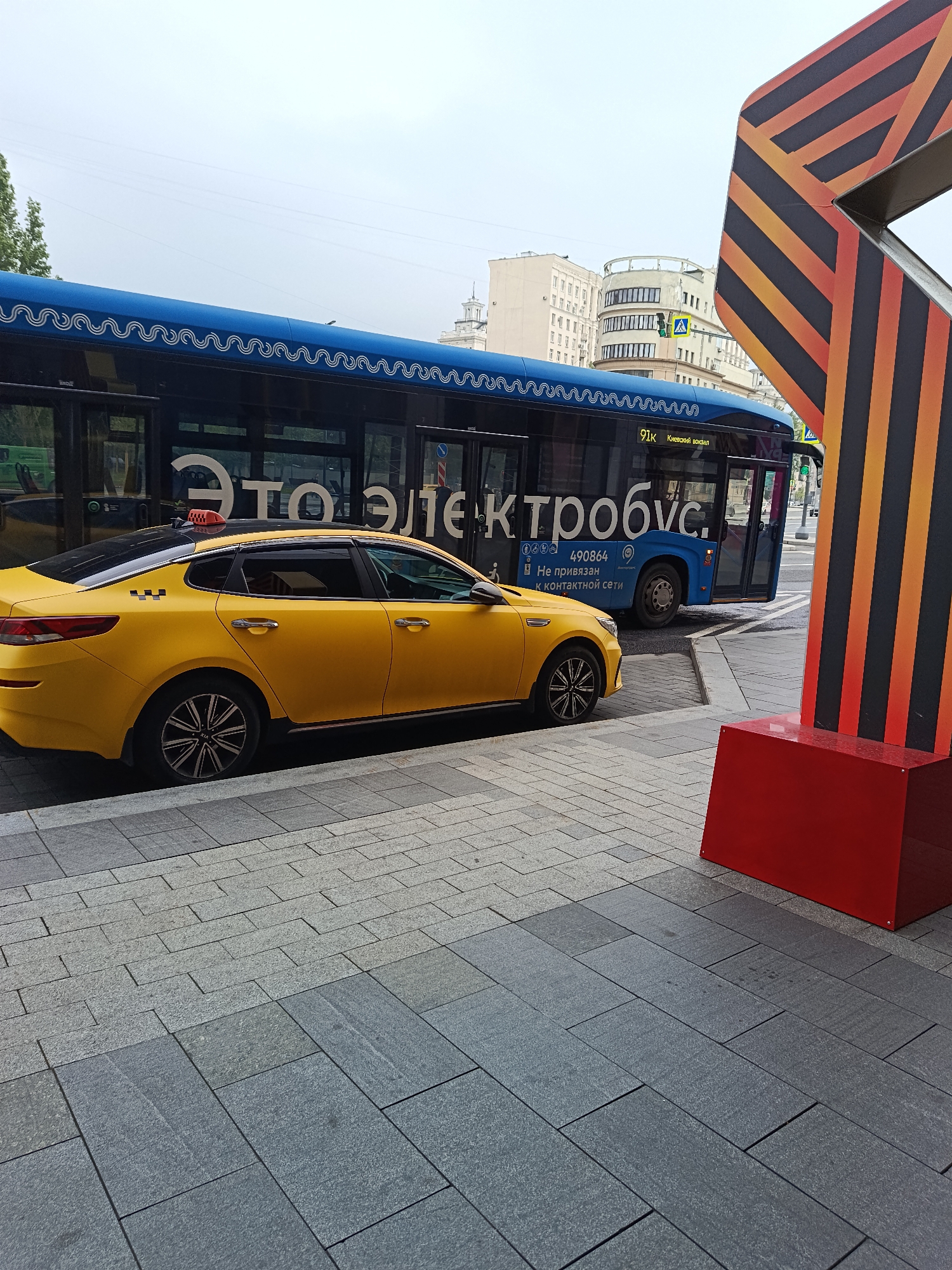 The image shows a yellow taxi parked on a grey stone-tiled sidewalk in Moscow, Russia. Adjacent to the taxi is a blue "Elektrobus" (electric bus) with large Cyrillic letters on its side, the bus route number "91K Киевский вокзал" and the words "Не привязан к контактной сети" and the number 490864 can be seen on the bus. To the right, a red and black striped structure stands prominently. The background includes a multi-story building with a tower-like structure, suggesting an urban environment. There is no person in the image. The general impression is of a regular city scene with public transportation and infrastructure.