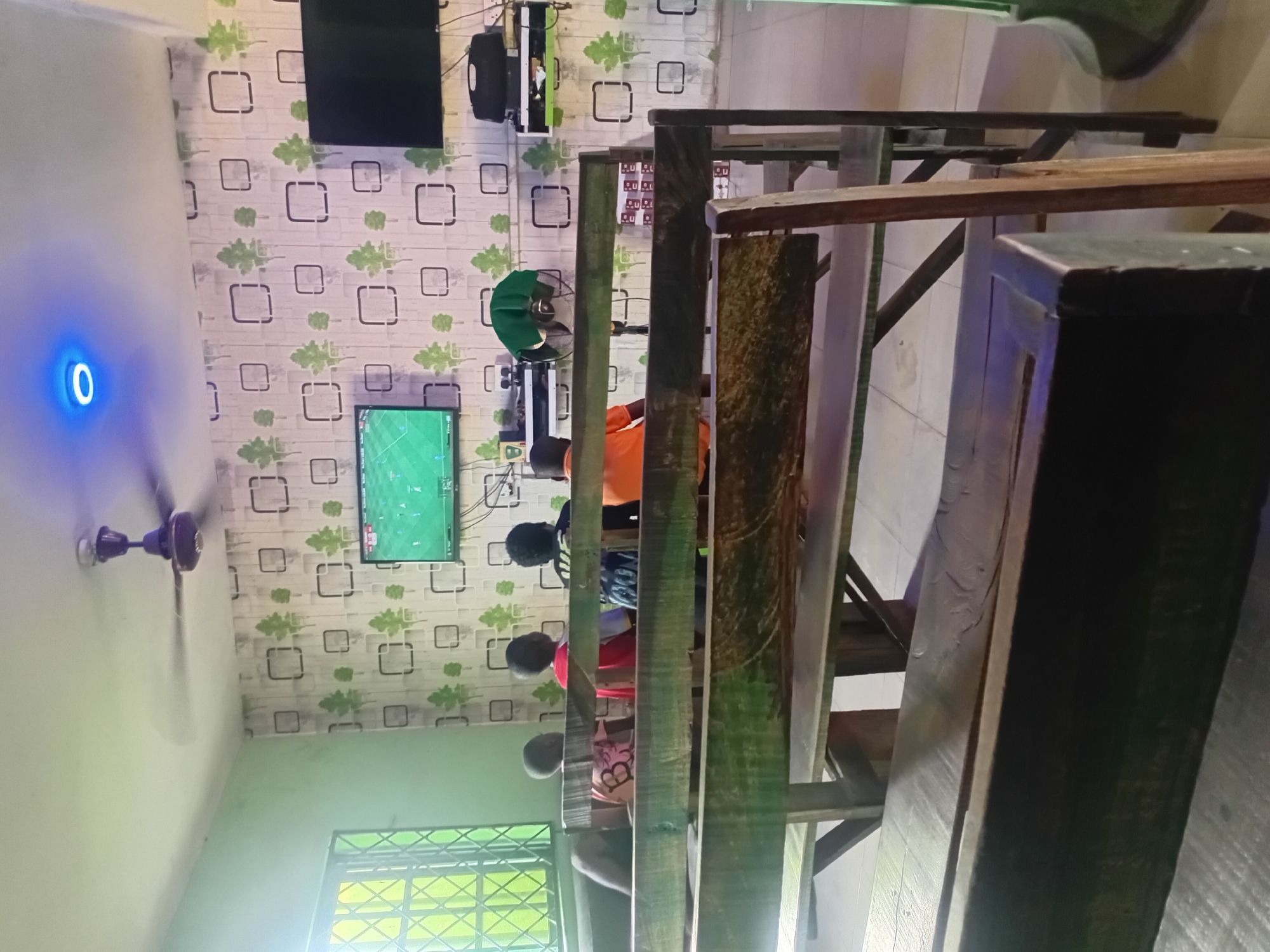 This image captures an indoor entertainment space in Lokoja, Nigeria, likely a gaming parlor or a communal viewing center. The scene is dominated by multiple wooden tables and benches, where several people, primarily children or young adults, are seated, mostly with their backs to the viewer.
The primary activity appears to be gaming or watching content on screens. A central television prominently displays a football (soccer) video game. Above it, another smaller screen is visible, and a larger, off-black television is mounted on the upper left wall. The area is equipped with cooling devices, including a standing fan with a green cover and a purple-brown ceiling fan on the left.
The walls are covered with distinctive wallpaper featuring a pattern of green leaf shapes and black square outlines. On the left wall, a round, blue glowing light fixture stands out, displaying a white numeral "0" in its center. A window with a green lattice pattern is visible on the bottom left. The wooden furniture has a rustic, unpolished appearance, suggesting a casual and possibly self-built or locally crafted aesthetic. Below the main gaming screen, various electronic devices, likely game consoles, are arranged on a shelf. The general lighting suggests it could be daytime or early evening. One person wears an orange shirt, another a red shirt, and others are in darker attire. On the back of one person's shirt, a faint, indistinguishable white letter or logo can be seen.