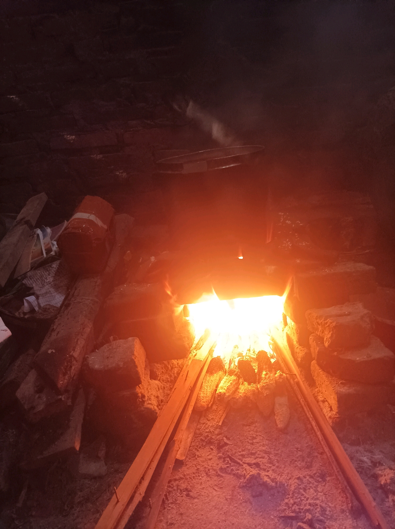 An indoor scene is depicted with a visible fire source and its surroundings. The fire is bright orange and white, casting a warm glow on the immediate area. The fire is contained within a brick structure, suggesting a furnace or hearth. Various objects are placed around the fire, including wooden planks, bricks, and what appears to be a metal pan or lid suspended above the flames. Some items on the left appear to be bags and cylindrical objects. The overall environment is dark, with the fire being the primary light source. The scene suggests a context of cooking or heating, possibly in a traditional setting.