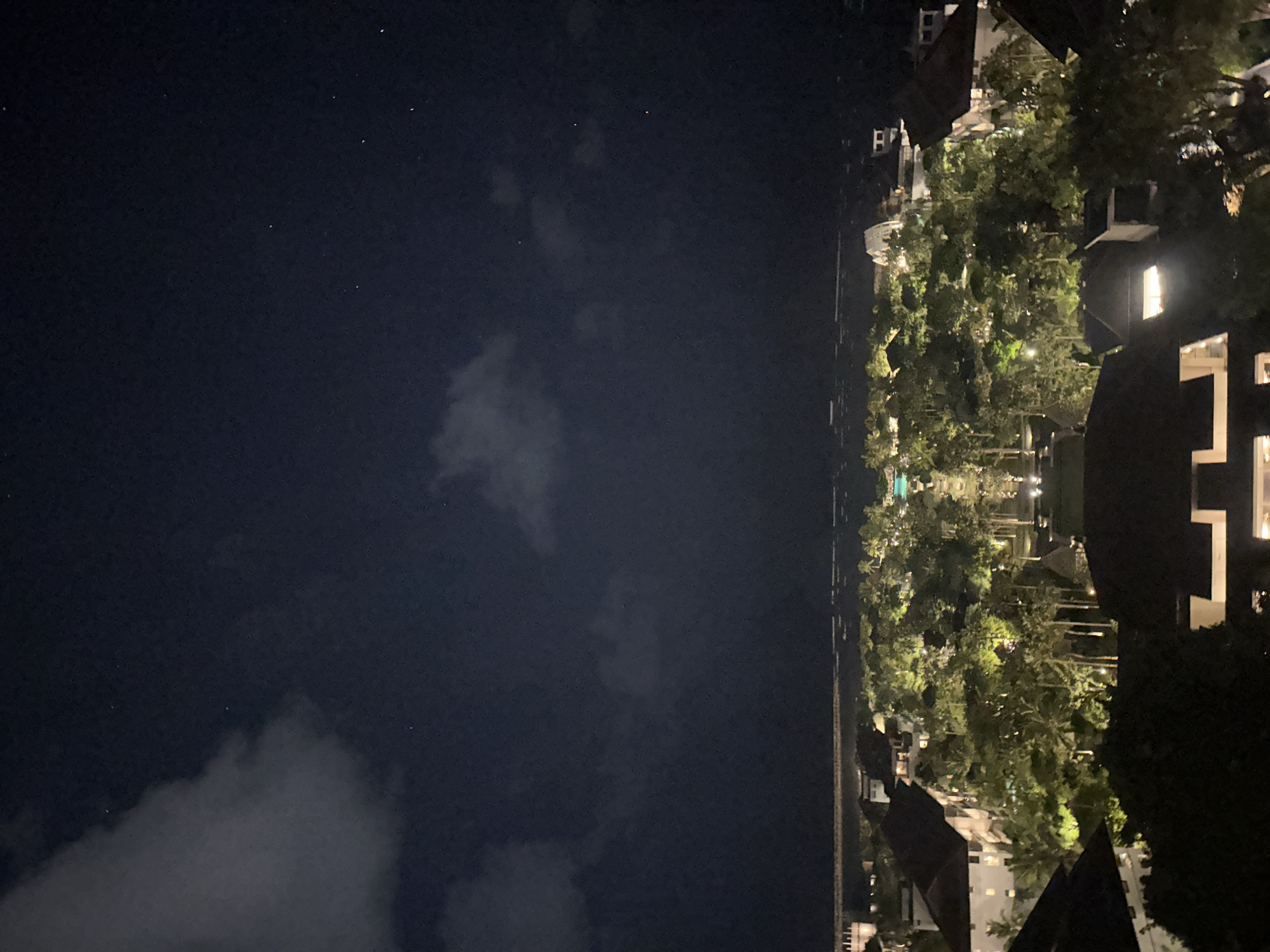 The image captures a serene nighttime scene over a resort or residential area in Kuta Selatan, Indonesia. The sky occupies roughly two-thirds of the frame, a deep navy blue canvas dotted with numerous faint stars. Scattered, wispy clouds are visible in the lower and central portions of the sky.
The remaining third of the image depicts a brightly lit landscape. Numerous lush trees, many appearing to be palm trees, are illuminated from below by artificial lights, casting a vibrant green glow. Interspersed among these trees are several buildings featuring white facades and dark, possibly tiled, roofs. Many windows in these structures are lit, suggesting they are occupied. A faint, horizontal line, likely a pathway or road, runs along the horizon, separating the illuminated ground from the dark sky.
No specific activity or people are visible, contributing to the tranquil and peaceful ambiance of a tropical night. The clear visibility of stars, despite some cloud cover, suggests minimal light pollution. There is no visible text in the image.