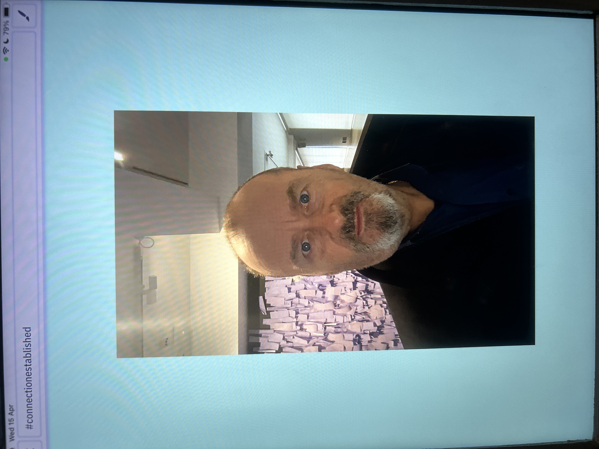 A photograph of a man with light skin, short hair, a beard, and blue eyes is displayed on an electronic device screen. The man is wearing a dark-colored shirt. Behind the man, there is a wall with a textured pattern that appears to be made of stacked, white rectangular blocks. The environment suggests an indoor setting. The device screen displaying the photograph has a light blue background. Text on the screen includes "#connectionestablished" and "Wed 15 Apr." The location is not ascertainable from the provided image.