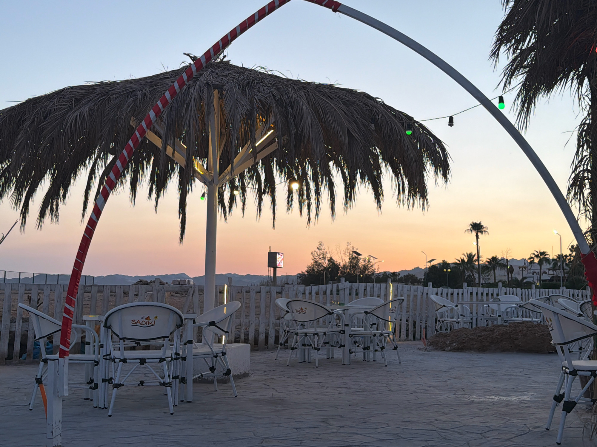 This image depicts an outdoor seating area at a beachside establishment in Sharm El Sheikh, Egypt, during twilight. The foreground features white metal tables and chairs, some with the "SADIKI" logo visible on their backs, arranged on a paved surface. A large palm frond umbrella provides shade, with decorative red and white striped poles supporting it. String lights, some emitting a green glow, are strung across the area and under the umbrella. In the background, a white picket fence separates the seating area from a sandy patch and further off, palm trees and distant hills are visible against a gradient sky transitioning from pale blue to orange and pink hues. A digital billboard is also visible in the mid-ground. The scene suggests a tranquil, possibly evening ambiance at a resort or restaurant.