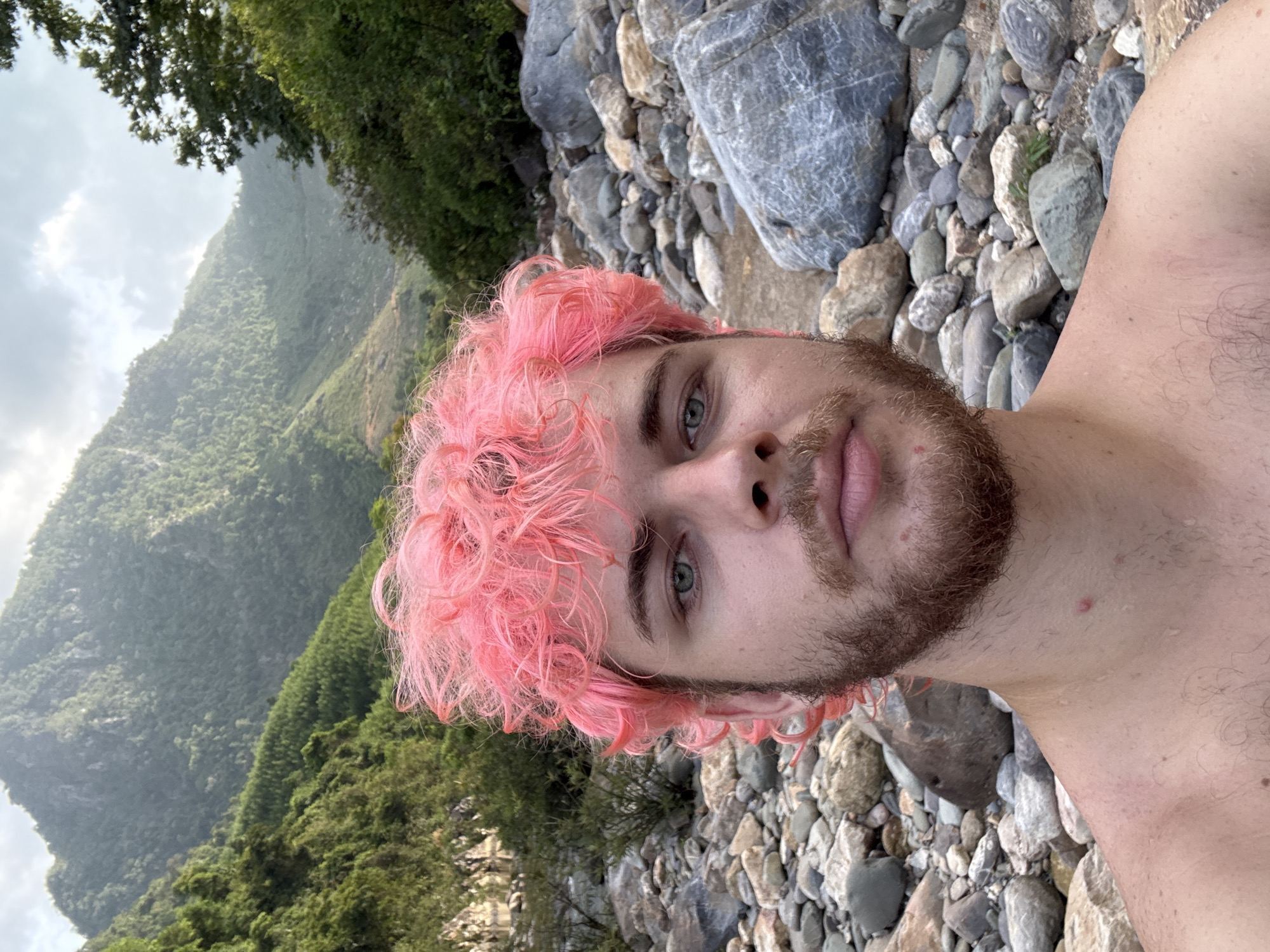 A selfie shows a fair-skinned person with pink curly hair and a beard, positioned against a backdrop of a mountainside covered in green vegetation and a foreground of rocks and pebbles. The person is shirtless, and their gaze is directed towards the camera. The setting appears to be a natural environment, possibly a riverbank or rocky terrain near a mountainous region. There are no indications of the city of Đà Nẵng, Vietnam, in the immediate scene.