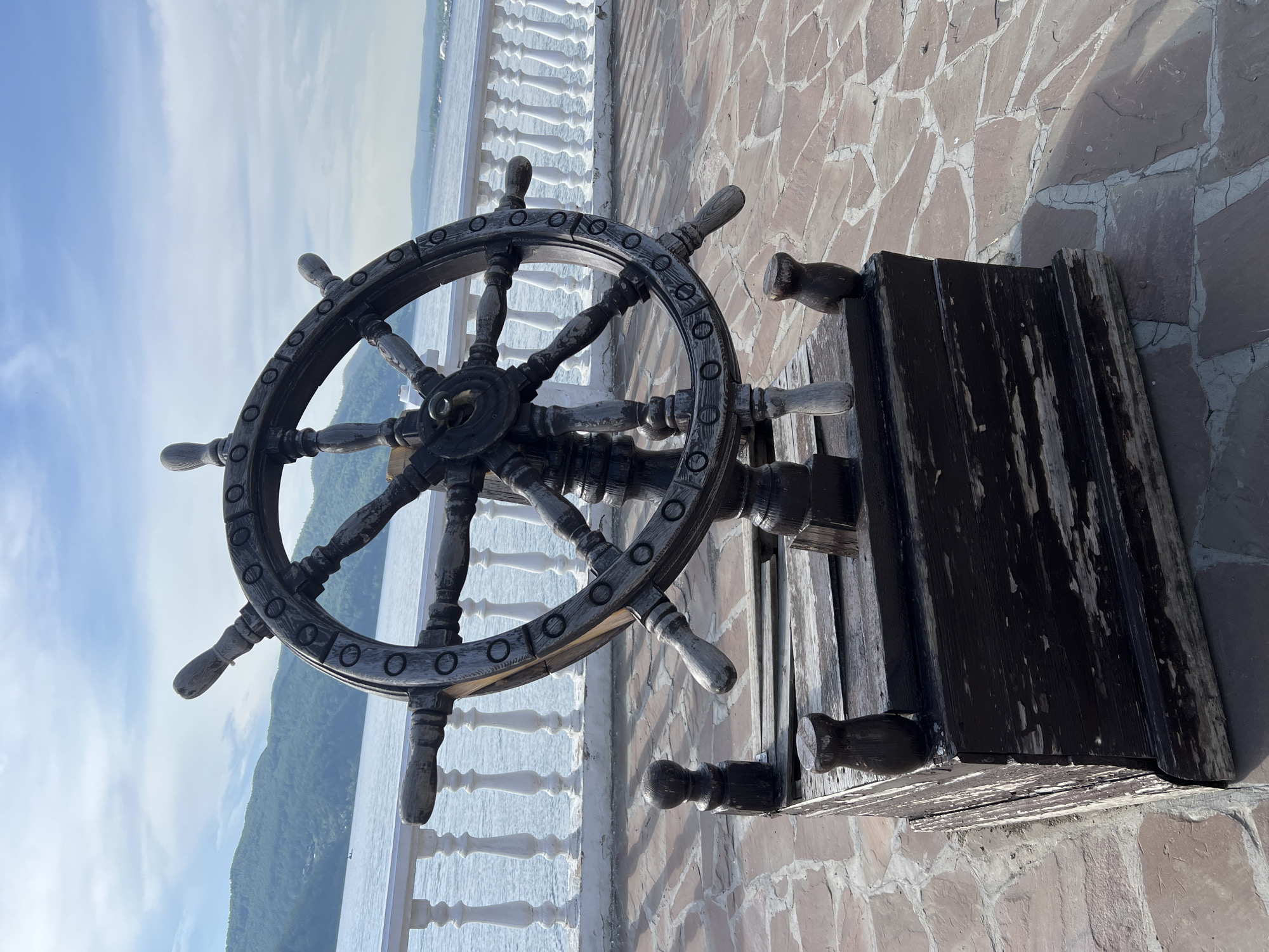 An antique, weathered wooden ship's steering wheel, ornately carved with circular details and mounted on a dark, distressed wooden base, stands prominently on a stone-paved walkway. In the background, a white ornate railing borders a large body of water, likely Lake Baikal, with lush, green, tree-covered hills rising in the distance under a bright, partially cloudy sky. The scene is set in Listvyanka, Russia, a popular tourist destination on the southwestern shore of Lake Baikal.