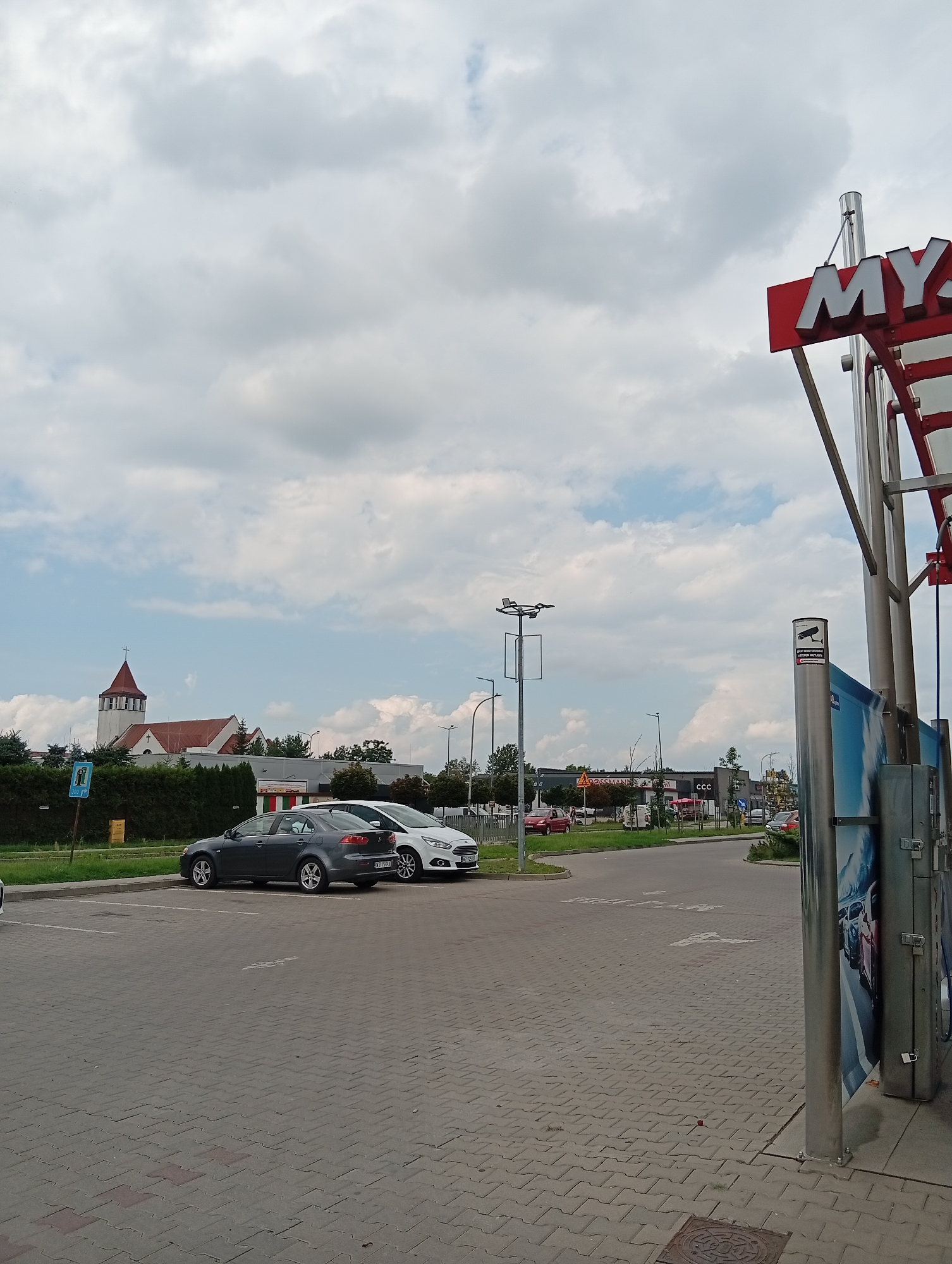 In Błonie, Poland, on a cloudy day, a car wash with a prominent red and white "MY" sign is visible on the right. Two cars, a dark gray sedan and a white hatchback, are parked in a paved lot in the foreground. In the background, a church with a red roof and a steeple stands to the left, and a row of businesses, including a "ROSSMANN" store, are situated along a road. The scene appears to be a commercial area.
