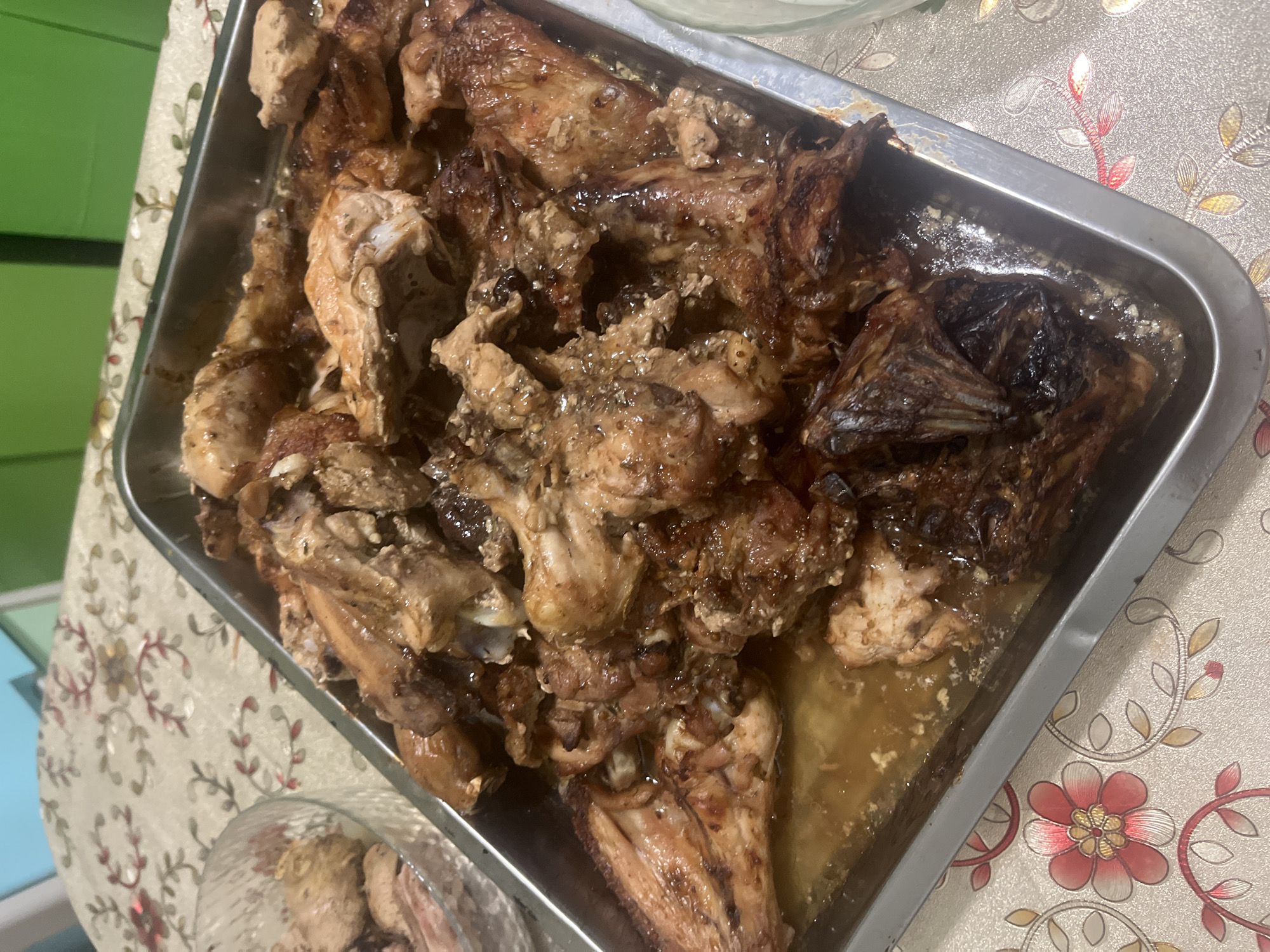 A close-up, high-angle shot shows a metal rectangular baking pan filled with cooked chicken pieces on a patterned tablecloth. The chicken is golden brown and appears to be glazed or sauced, with some pieces showing charring. The pan is brimming with chicken, making it difficult to discern individual pieces clearly. The tablecloth has a floral design with pink and red flowers and green leaves on a light beige background. In the upper left corner, a glass bowl containing more cooked chicken is partially visible. The background is slightly out of focus, showing a greenish surface and a hint of another object. The scene appears to be set indoors, likely in a dining area. There is no indication of the city of Tolsty, Russia, or any specific environmental details related to its location.