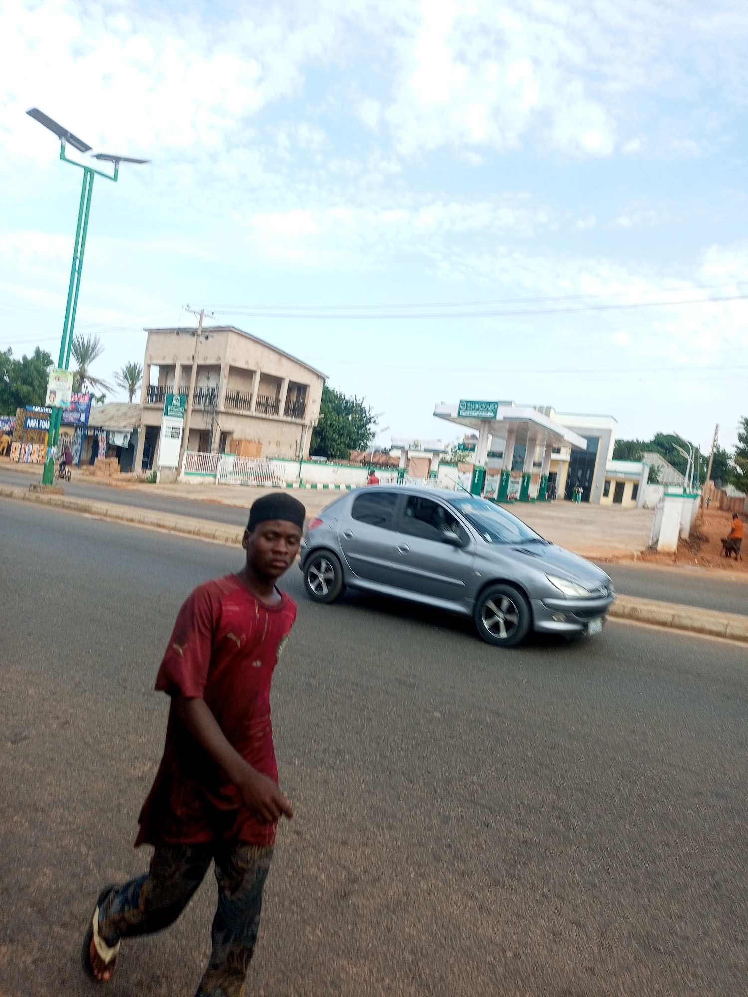 In the city of Gombe, Nigeria, a young male wearing a maroon t-shirt and patterned trousers walks on an asphalt road. He is looking down and to his right. Behind him, a silver Peugeot 206 drives on the road. Further in the background, a gas station with green and white pumps is visible, along with a building with multiple balconies and a street lamp with solar panels. The sky is overcast with clouds.