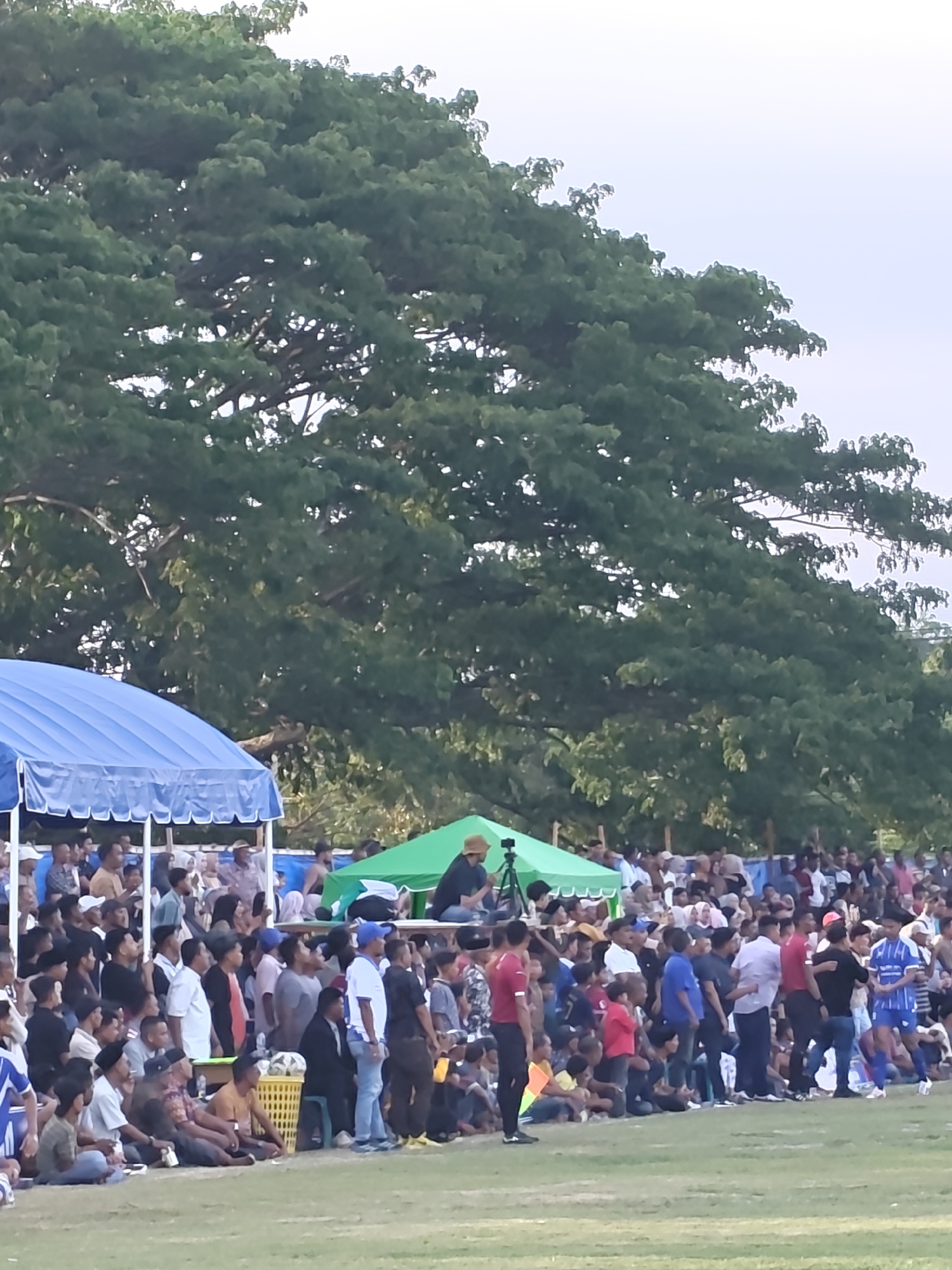 A wide shot captures a large crowd gathered on a grassy field in Desa Meunasah Blang, Indonesia, under a canopy of lush green trees. The spectators are arranged in tiers, with some sitting on the ground and others on elevated platforms or benches. Two tents, one blue and one green, are set up near the crowd, providing shade. Several people are visible in the foreground, some wearing athletic attire, suggesting a sporting event is taking place. The overall atmosphere appears to be one of community engagement and participation in an outdoor activity.