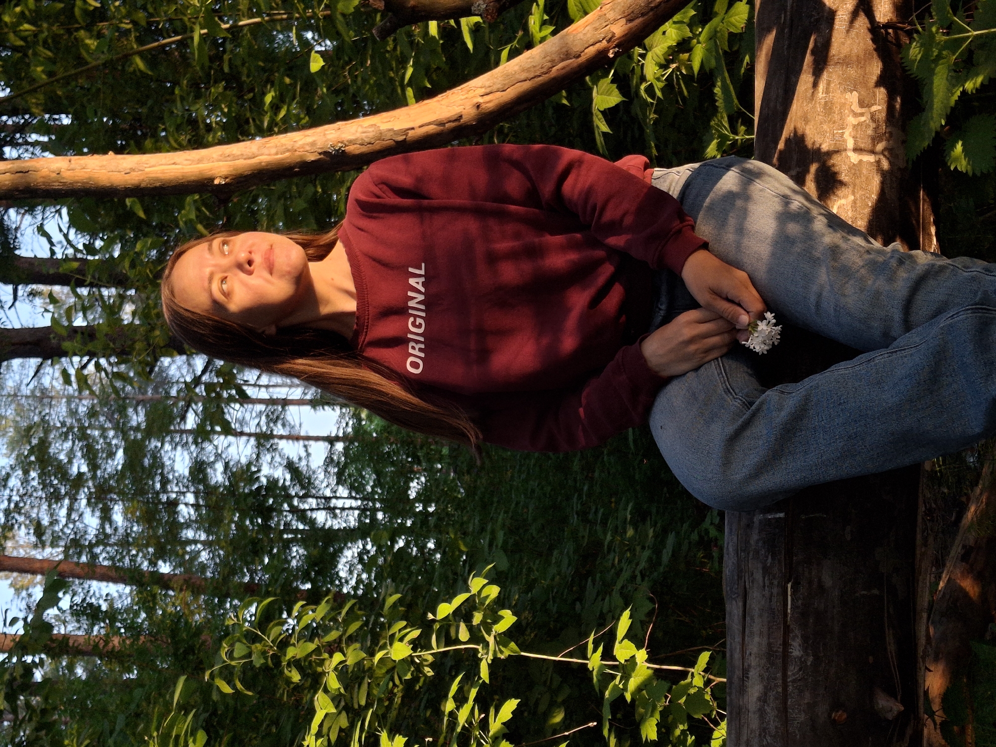 The image shows a woman lying on a fallen tree trunk in a forest setting. She is wearing a maroon sweatshirt with the word "ORIGINAL" printed vertically on the front and blue jeans. She holds a small bunch of white flowers in her hands. Sunlight filters through the trees, casting shadows on the ground and on the woman's face. The location is Chaikovsky, Russia.
