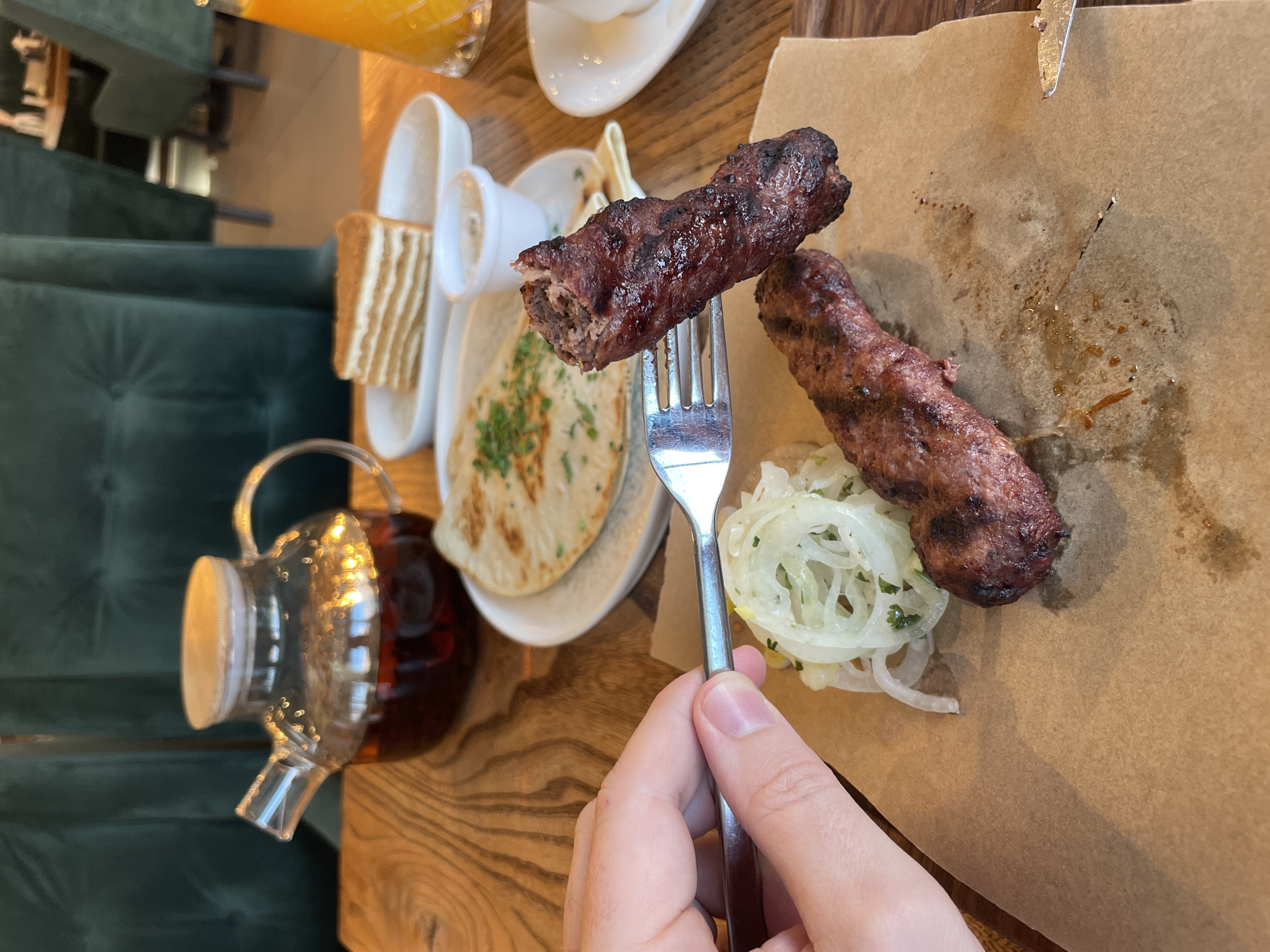 A first-person perspective shot shows a hand holding a fork with a piece of grilled meat on it, hovering over a plate of food. The plate contains a piece of grilled meat, thinly sliced onions with herbs, and what appears to be flatbread or naan bread in the background. In the background, a glass teapot filled with dark liquid sits on a wooden table, alongside a slice of honey cake and a glass of orange juice. The scene is set in what appears to be a restaurant with green velvet chairs visible. The overall lighting suggests an indoor setting.