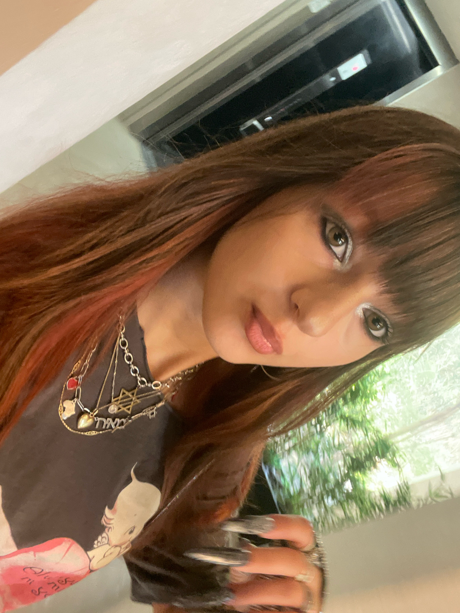 A young woman with long, reddish-brown hair and bangs is taking a selfie in Raanana, Israel. She is wearing a dark t-shirt with a cartoon image on it and several necklaces, including one with a Star of David pendant. Her eyes are adorned with silver eyeshadow, and her lips are painted with a nude lipstick. The background shows a blurry image of greenery through a window and part of a light-colored wall. The image is tilted, suggesting it was taken at an angle.