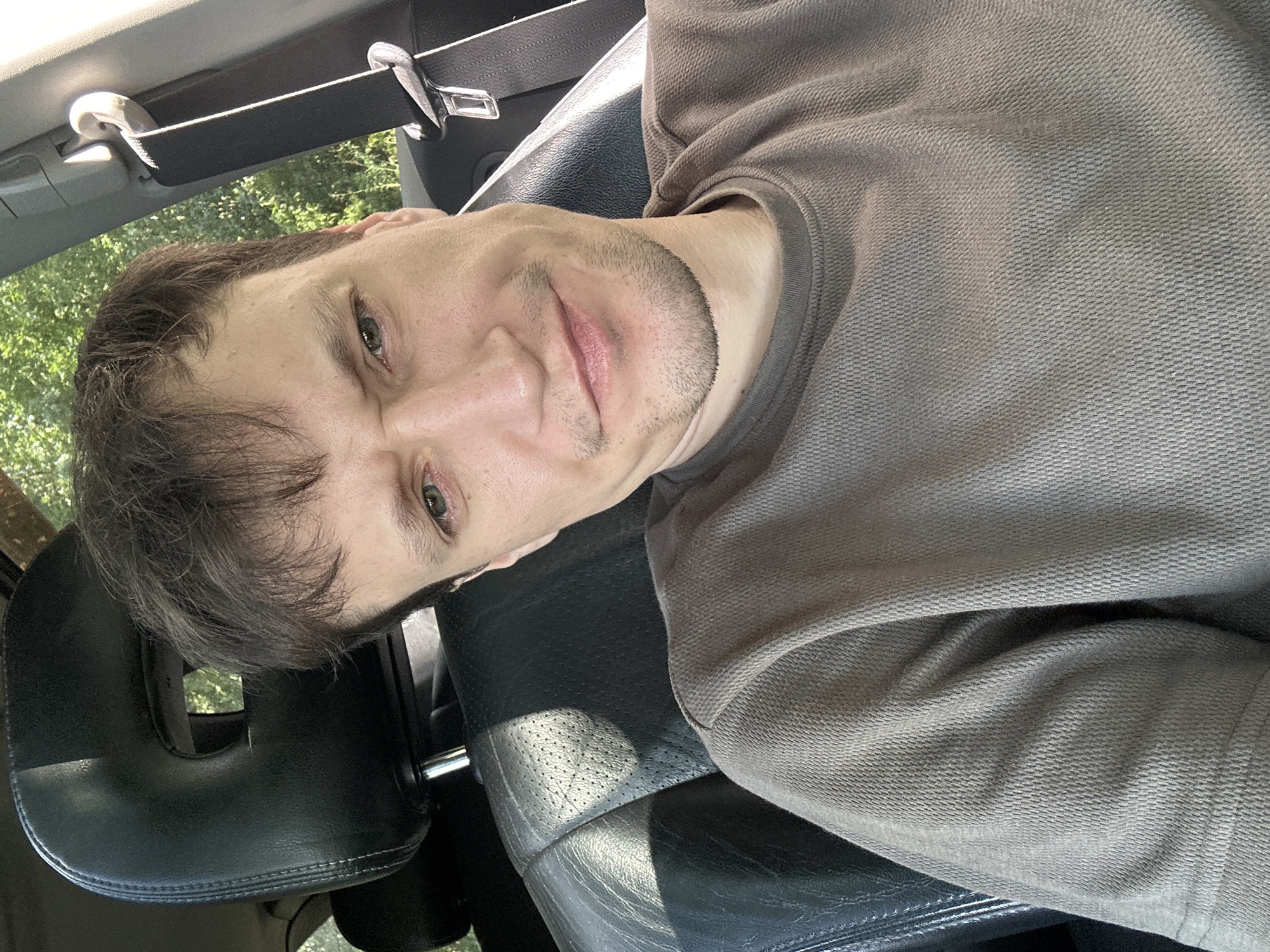 A middle-aged man with short, brown hair and a light stubble is seated in the driver's seat of a car. He is looking directly at the camera with a slightly furrowed brow and a subtle smile. He is wearing a brown, textured crew-neck shirt. The interior of the car is visible, including a black leather headrest and seat, and a seatbelt buckle. Sunlight streams in from the right, illuminating the scene. Outside the car, green foliage suggests an outdoor, possibly rural or suburban, environment. The image appears to be a selfie taken by the man. The location in the city of Shatava, Ukraine is not discernible from the image content.