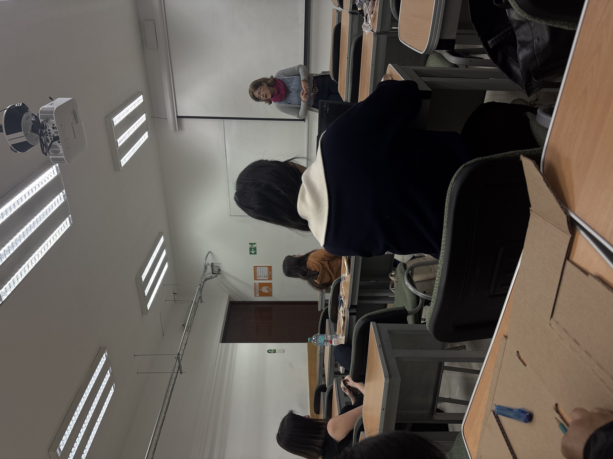 The image depicts a classroom scene at a university in San Andrés Cholula, Mexico. A female instructor stands at the front of the room by a projector screen. A class of students sits at desks, facing the instructor. Fluorescent lights illuminate the room. The furniture consists of individual desks and chairs. The atmosphere seems academic and focused. Some students engage with materials on their desks. There are no external visible landmarks in this image to identify exact location details.