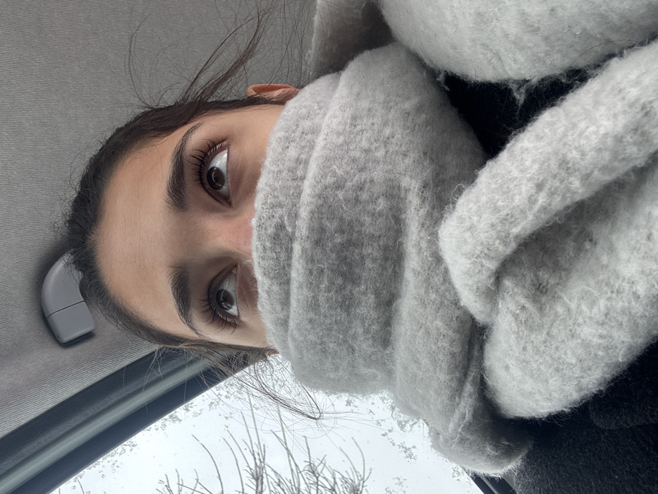 The image displays a close-up, upward-angled interior view of a vehicle, focusing on an individual from the mid-nose upwards. The subject has dark eyes, defined eyebrows, and prominent eyelashes, with their gaze directed upwards. A thick, light grey, textured scarf envelops the lower portion of the face, covering the mouth and chin. Dark hair strands are visible around the temples. The vehicle's grey fabric headliner is visible above the subject's head, along with a grey plastic overhead handle or clip on the left. Through a window on the upper left, an outdoor environment is discernible, characterized by snow or frost on visible surfaces and bare tree branches against a light, overcast sky, indicating winter conditions. The image provides no visual information to corroborate the specified location as Cibukovac, Serbia.