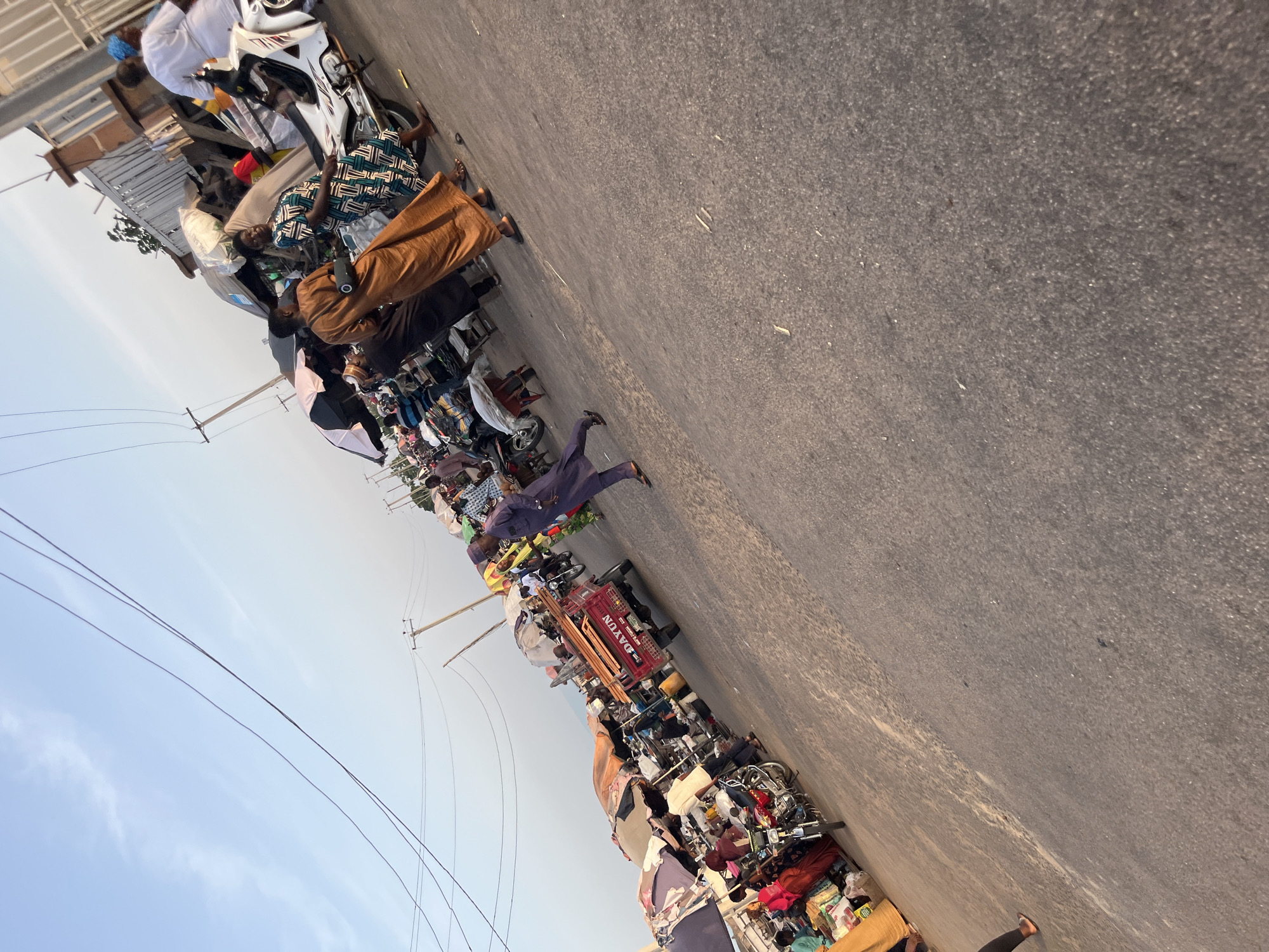 The image depicts a busy outdoor market scene. Several individuals are present, dressed in a mix of traditional and modern attire. Some people are walking, while others appear to be browsing or interacting with vendors. There are numerous stalls and makeshift shops, many covered with colorful umbrellas, displaying a variety of goods, including textiles and what appear to be food items or household products. Motorcycles and a small red truck are also visible, suggesting modes of transportation and potential delivery services within the market. The ground is paved, and utility poles with electrical wires are present in the background, indicating an urban or semi-urban setting. The overall atmosphere appears to be one of lively commerce and community interaction.