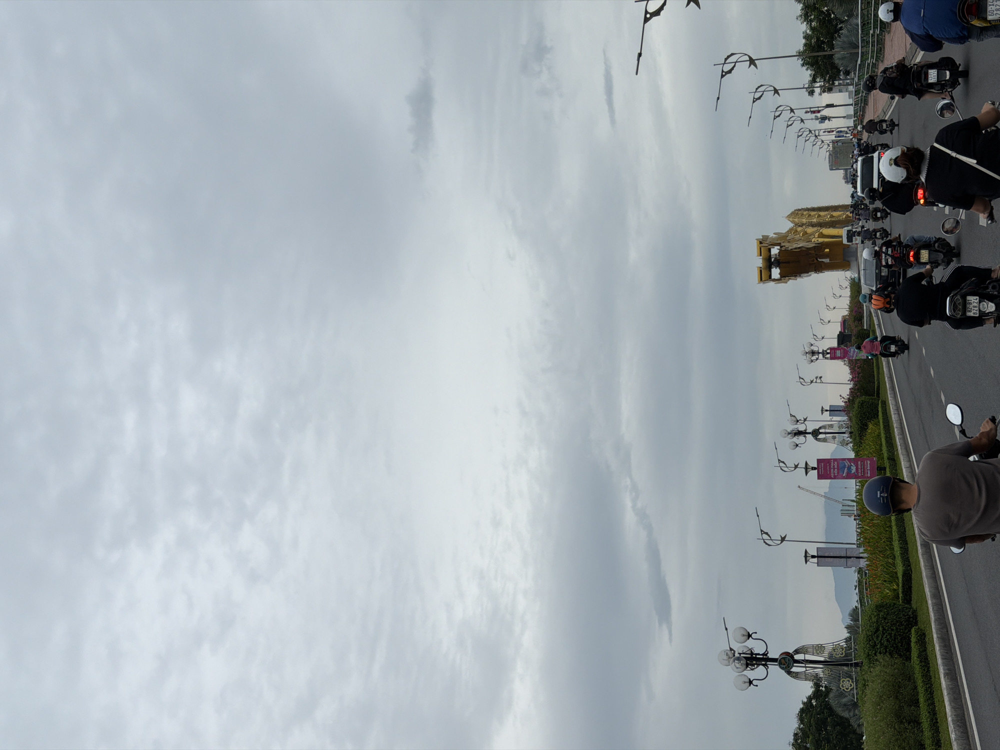 The image shows a street scene in Da Nang, Vietnam, likely on Dragon Bridge, given the prominent golden dragon-shaped structure visible in the background. The sky is overcast with a significant amount of clouds. The street is paved and marked with lane dividers, with multiple lanes of traffic. A procession of motorbikes, with riders wearing helmets, is moving along the road. Several cars are also present in the traffic. On the sides of the road, there are decorative streetlights with multiple bulbs, lush green vegetation, and manicured hedges. There are also billboards and signs visible along the route. The overall impression is of a busy urban environment on a cloudy day.