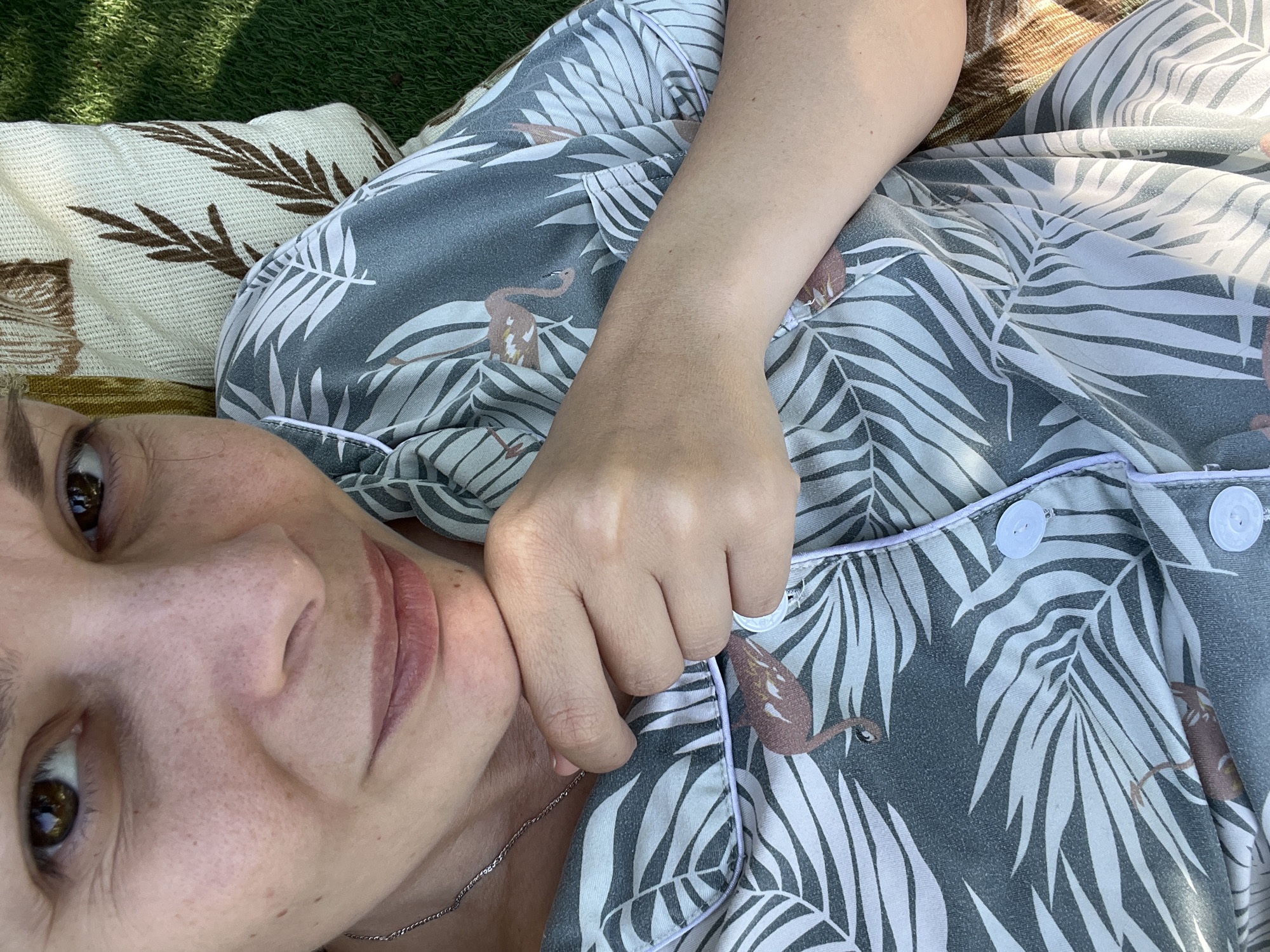 The image is a close-up, selfie-style shot of a woman's face and upper body, taken outdoors in Khryashchevka, Russia. The woman is lying down, with her head propped on a pillow that has a brown leaf pattern, situated on grass. She is wearing a gray pajama top with a white palm leaf and flamingo print. Her right hand is resting on her chin, and she is wearing a ring on her index finger. The lighting suggests it is daytime, with sunlight casting shadows.