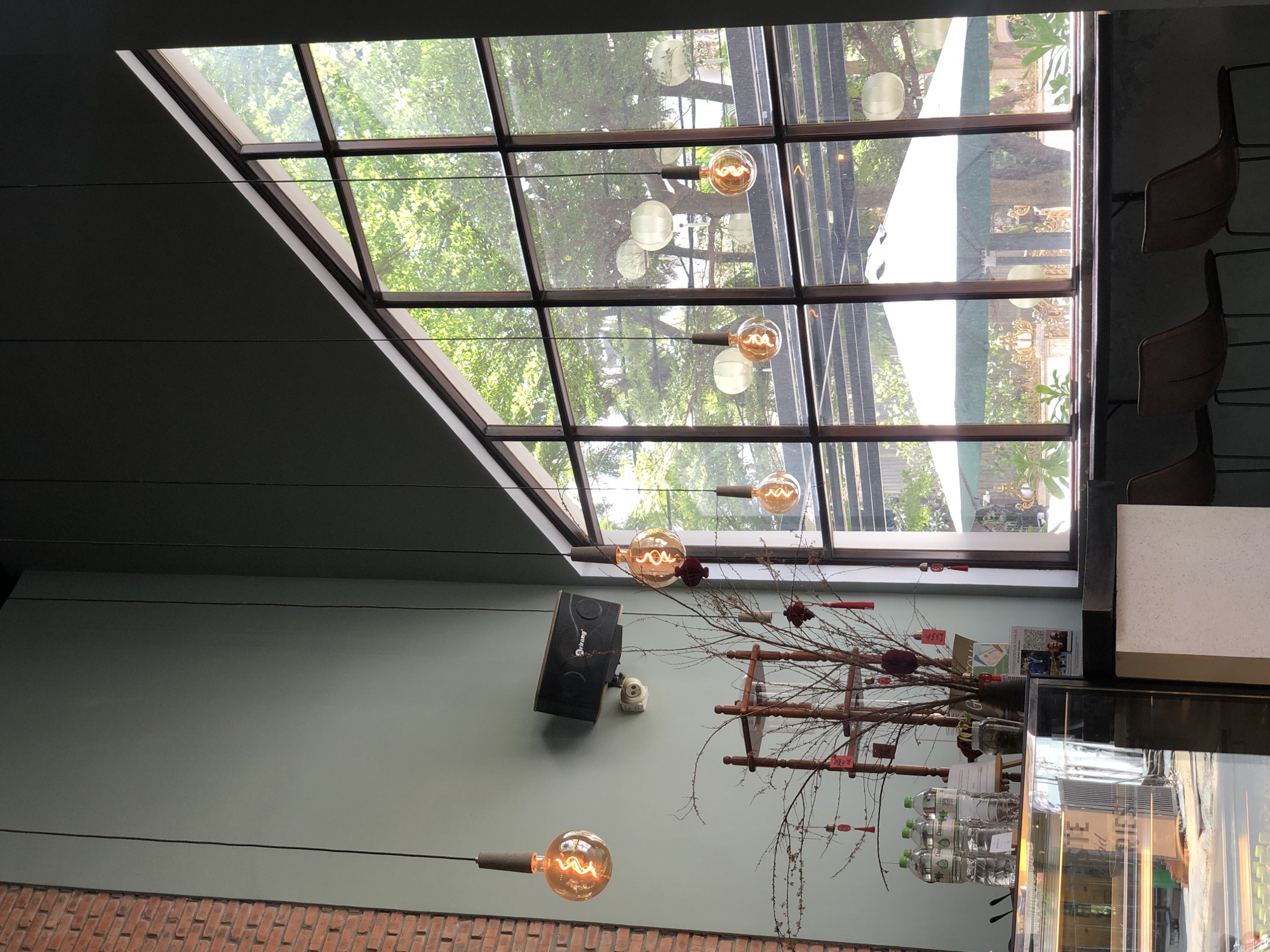This image depicts the interior of a cafe in Đà Nẵng, Vietnam, characterized by its high ceilings and large, angular windows offering a view of lush greenery and outdoor seating. The interior features exposed brick on the left wall, contrasting with a muted green painted wall. Several Edison-style filament bulbs hang from the ceiling, casting a warm glow. A black speaker is mounted on the green wall above a decorative tree branch adorned with red ornaments and small tags. To the left, a glass display case is visible, holding various items. The large window is divided into multiple panes, revealing an outdoor seating area with large white umbrellas and decorative outdoor furniture. The overall ambiance suggests a modern yet cozy cafe setting.