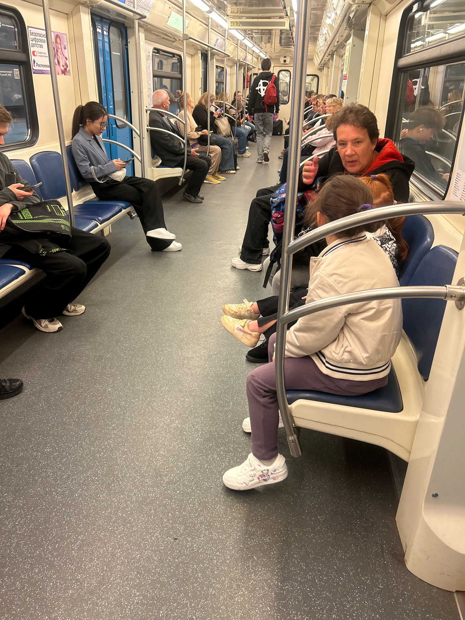 The image depicts the interior of a Moscow Metro train car. Several passengers are seated and standing. In the foreground, a young woman with dark hair pulled back in a ponytail, wearing glasses, a blue blazer, and black pants, is seated and looking at her phone. Next to her, a person in dark clothing is seated. Further down the aisle, a woman with short, dark hair, wearing a red and black hoodie, is speaking while looking at another person. A young girl with red hair is seated in front of her. Other passengers are visible throughout the car, some looking at their phones, others conversing. The train car has blue seats and a grey, speckled floor. Overhead lights illuminate the interior. The scene suggests a typical commute on public transportation in Moscow.