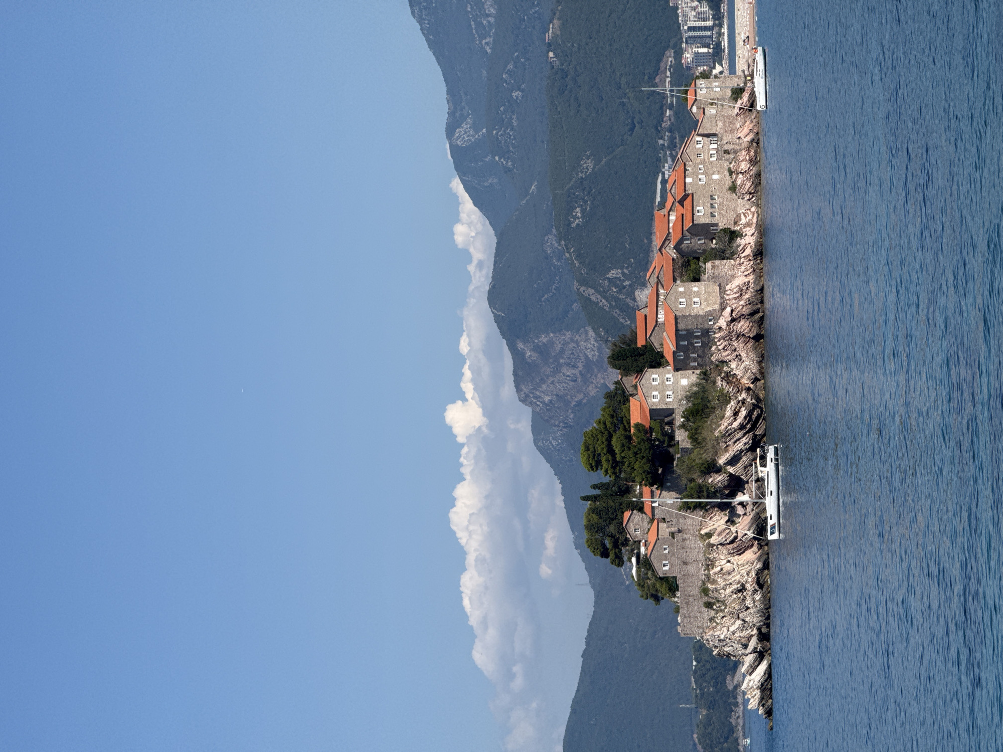 A vertical shot captures the Sveti Stefan island in Montenegro, jutting out into the calm blue Adriatic Sea under a clear sky with scattered white clouds. The island is densely packed with old stone buildings topped with red-tiled roofs, nestled atop rocky cliffs. Green trees grow amidst the structures. A white sailboat with a tall mast is anchored in the water near the island, with another smaller boat visible further in the distance. The background features a chain of lush green mountains, and the hazy outlines of a distant cityscape can be seen on the right. The water exhibits a rippled texture from a gentle breeze.