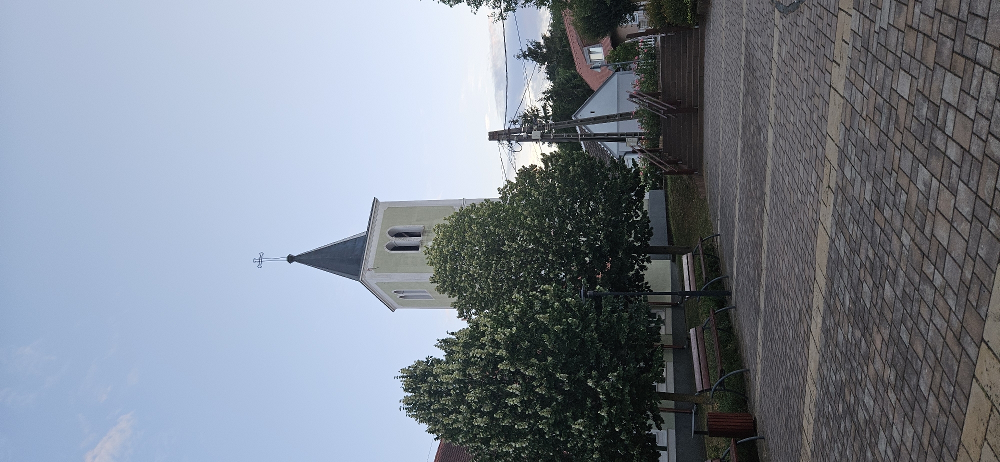 The image captures an outdoor scene in Hévíz, Hungary, featuring a church steeple prominently in the background. The steeple is tall and slender, topped with a cross. It is a light yellow color with a dark grey, conical roof. Adjacent to the steeple, to the left, are two large, leafy trees. In front of the trees, there are several park benches, some appearing empty. The foreground is a paved area with a patterned cobblestone surface. To the right of the church steeple, a set of outdoor stairs with railings leads upwards towards buildings with red-tiled roofs. Electrical poles and wires are visible behind the trees and near the stairs, indicating the presence of infrastructure. The sky is clear and pale blue, suggesting either early morning or late evening light. The overall scene conveys a peaceful, somewhat idyllic atmosphere of a small town or village setting.