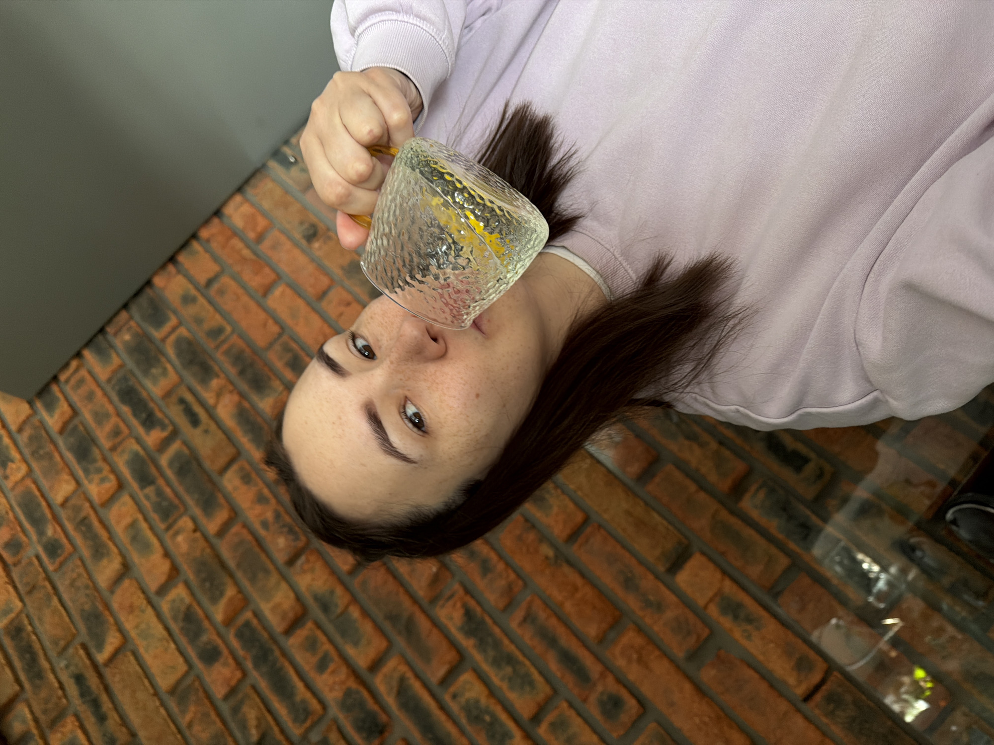 A high-angle shot captures a person lying down or significantly tilted, looking directly upwards towards the camera. The image itself is rotated 90 degrees clockwise, positioning the person's head on the left side of the frame and their body extending to the right. The individual has fair skin with noticeable freckles across their nose and cheeks, dark brown hair, and brown eyes. They are wearing a light lavender or pale purple long-sleeved sweatshirt.
The person holds a clear, textured glass mug with a golden-yellow handle to their mouth, appearing to drink or about to take a sip. The mug contains a clear liquid, possibly water, which reflects light. Their expression is neutral.
The background is divided into two main areas. Below and to the right of the person, a floor or wall covered in reddish-brown and dark-gray bricks in a stretcher bond pattern creates a rustic texture. To the upper left, a plain, smooth gray wall is visible. The lighting is soft and even, indicative of an indoor setting. No visible text is present. The overall scene suggests a casual, perhaps candid, moment.