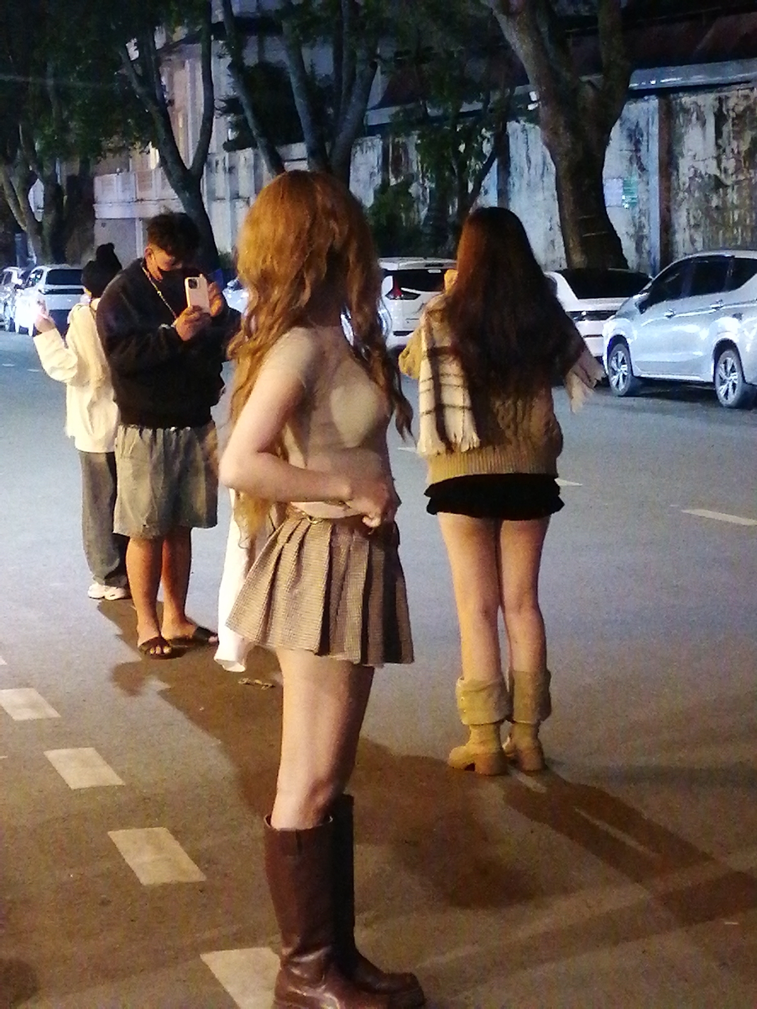 In Da Lat, Vietnam, a group of four individuals stands on a paved street at night. A person in the foreground, with long, wavy auburn hair, faces away from the camera, wearing a light-colored, sheer top and a patterned pleated skirt, paired with dark brown boots. To their left, a man wearing a black hooded sweatshirt, a face mask, and a necklace is looking at his phone. He is wearing grey shorts and brown sandals. Next to him, a person in a white long-sleeved shirt and grey pants stands with their phone in hand. On the right, another individual, seen from the back, wears a tan cable-knit sweater with a checkered scarf, a black skirt, and light brown boots. The street is lined with parked cars, including silver sedans and SUVs, and illuminated by streetlights, casting long shadows on the asphalt. Trees with dense foliage are visible behind the individuals, and a textured wall with peeling paint forms the background.