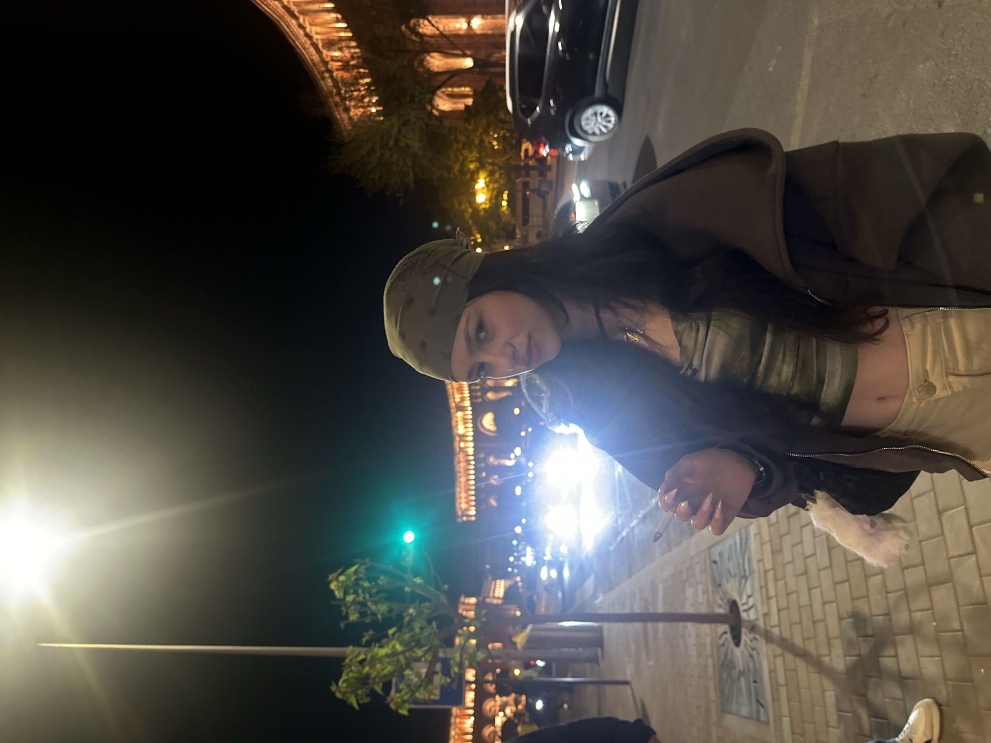 The image shows a woman standing on a brick sidewalk in Yerevan, Armenia, at night. She is wearing a green bandana, a green tube top, a dark jacket, and light-colored pants. She is holding a lit cigarette in her right hand. Behind her is a streetlight and a line of cars with their headlights on. In the background is a building with illuminated arches.