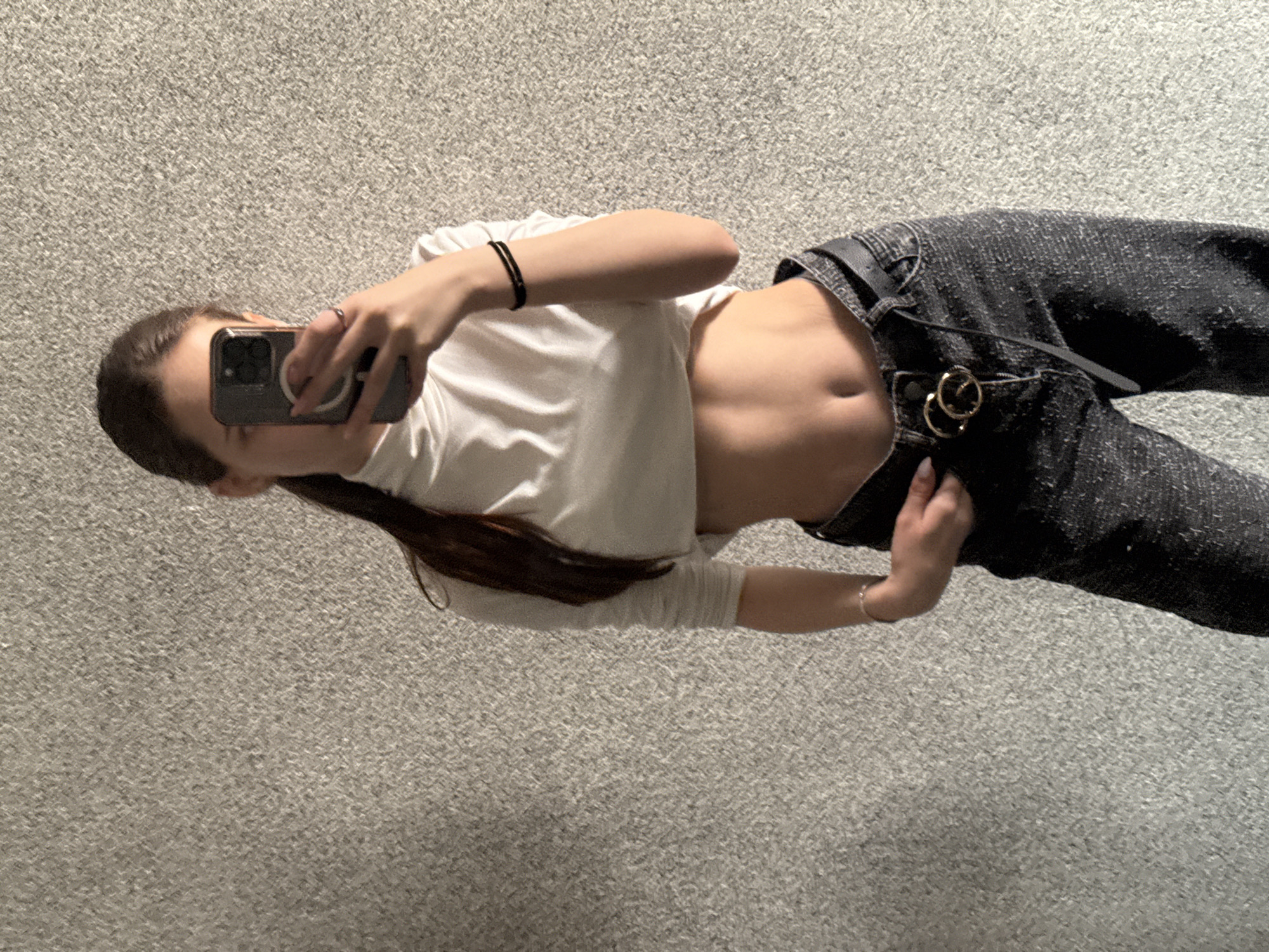 A female is lying on a gray carpet while taking a selfie with an iPhone. She is wearing a short white t-shirt, which reveals her midriff, and dark jeans with a black belt. Her dark hair is long. The location is unknown, and there is no evidence to suggest it is Sofia, Bulgaria.