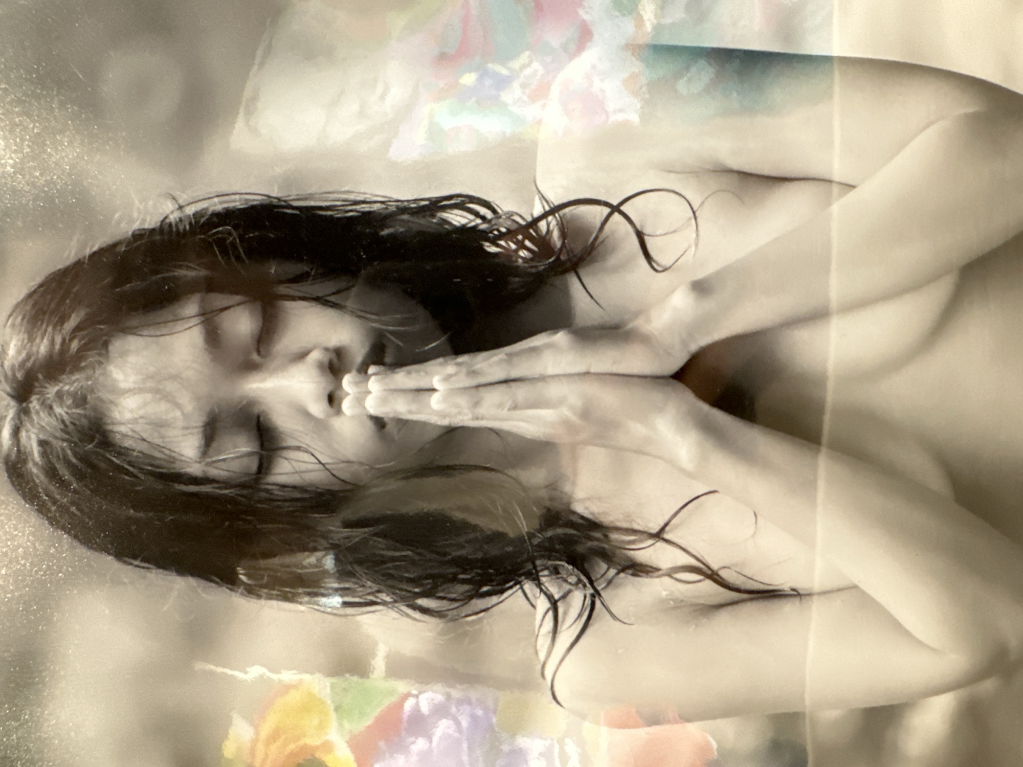 A topless Asian woman with wet, dark hair pulled back from her face, stands with her eyes closed and hands pressed together in a prayer-like gesture. She is positioned in the center of the frame. Abstract, colorful shapes are overlaid on the left and right sides of the image, suggesting a layered or artistic presentation. The background is a textured, light brown color with speckled highlights. The overall tone of the image is soft and somewhat ethereal. No specific location in Kuta, Indonesia, can be identified from the image itself.
