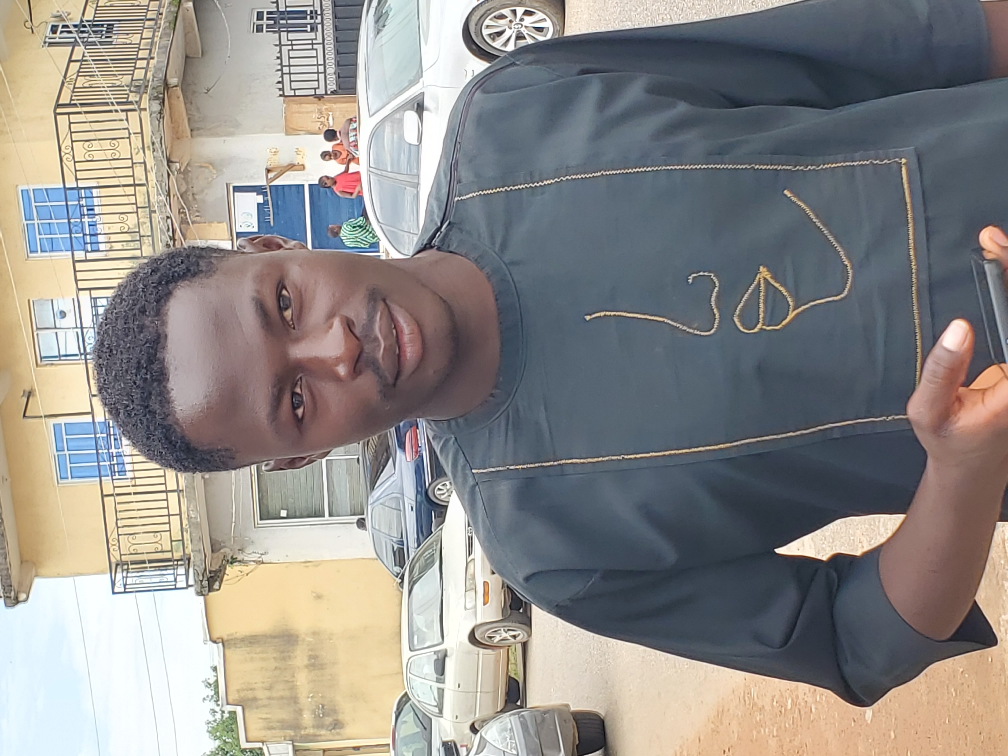This image depicts a dark-skinned male subject, approximately 20-35 years old, positioned centrally in the foreground, making direct eye contact with the camera. He has short, dark, tightly curled hair, a neatly trimmed mustache, and light facial stubble. He wears a dark-colored (likely black or navy) long-sleeved tunic, possibly a traditional garment, featuring gold-colored embroidery. The embroidery on the chest forms an abstract, minimalist profile of a face, specifically outlining a nose and lips. His left hand holds a dark-colored smartphone, partially visible near his lower chest.
The scene is set outdoors on what appears to be a street in the city of Jos, Nigeria. In the midground, several vehicles are parked. A light-colored (beige or white) Toyota Corolla sedan is prominent on the left, and a dark blue sedan (possibly a Peugeot) is visible behind it to the right. Beyond the vehicles, a group of four individuals is standing or conversing near a building entrance. One person on the far left of this group is wearing a green and white striped shirt, while another in the center wears a red top.
The background is dominated by a multi-story building with light yellow or beige exterior walls and prominent blue-tinted windows. The second floor features a black metal balcony railing with decorative scrollwork. The architecture suggests a common building style in urban Nigerian settings. The sky above is bright and overcast, indicating natural light. The ground in the immediate foreground is an unpaved reddish-brown surface, transitioning to a paved road further back where the cars are parked. Sparse green foliage is visible on the far left.