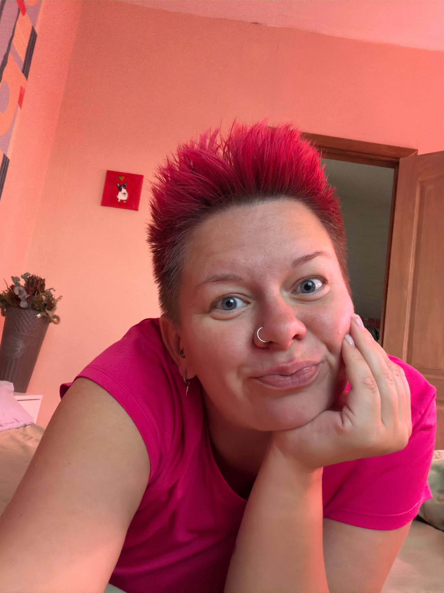 The image shows a close-up selfie of a woman with bright pink, spiky hair, blue eyes, and a silver nose ring. She is wearing a bright pink t-shirt and has her hand resting on her chin, looking at the camera with a playful expression. In the background, the wall is painted a peachy-pink color. To the left, there is a small framed picture of a black and white dog with a heart above its head, and to the right of that, a grey pot with dried flowers. A doorway with a light-colored door is visible in the upper right corner. The environment suggests an indoor setting, likely a bedroom, given the presence of a bed in the lower left corner of the frame. The lighting is warm and appears to be artificial. The image's content does not provide any specific information about the location within the city of Chaikovsky, Russia.