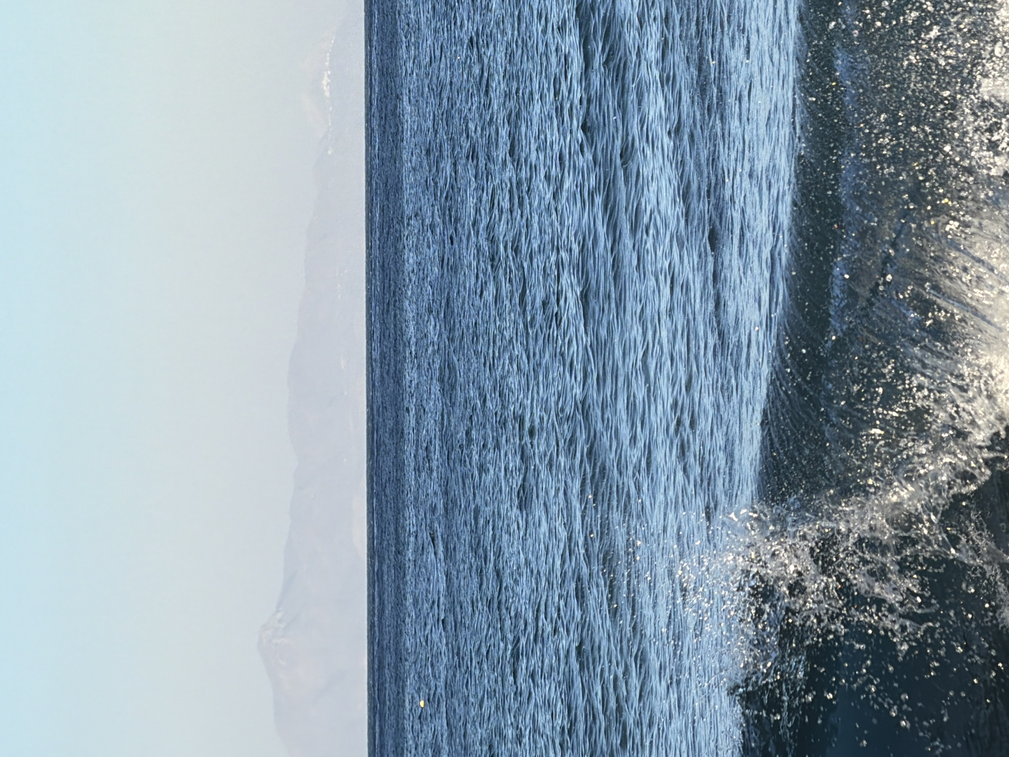 The image depicts a view of the Aegean Sea, with a clear sky and distant mountains. The water is deep blue and appears calm, with small waves and some whitecaps in the foreground. The horizon line is sharp, separating the sea from the sky. In the far distance, hazy mountain ranges are visible, with a faint suggestion of human settlements along the coastline. The scene appears to be a natural landscape, likely in the Thira Municipal Unit of Greece, given the characteristic volcanic topography of the region and the clear, azure waters of the Aegean. There are no people or man-made structures immediately visible in the foreground or mid-ground. The overall impression is one of serene natural beauty.