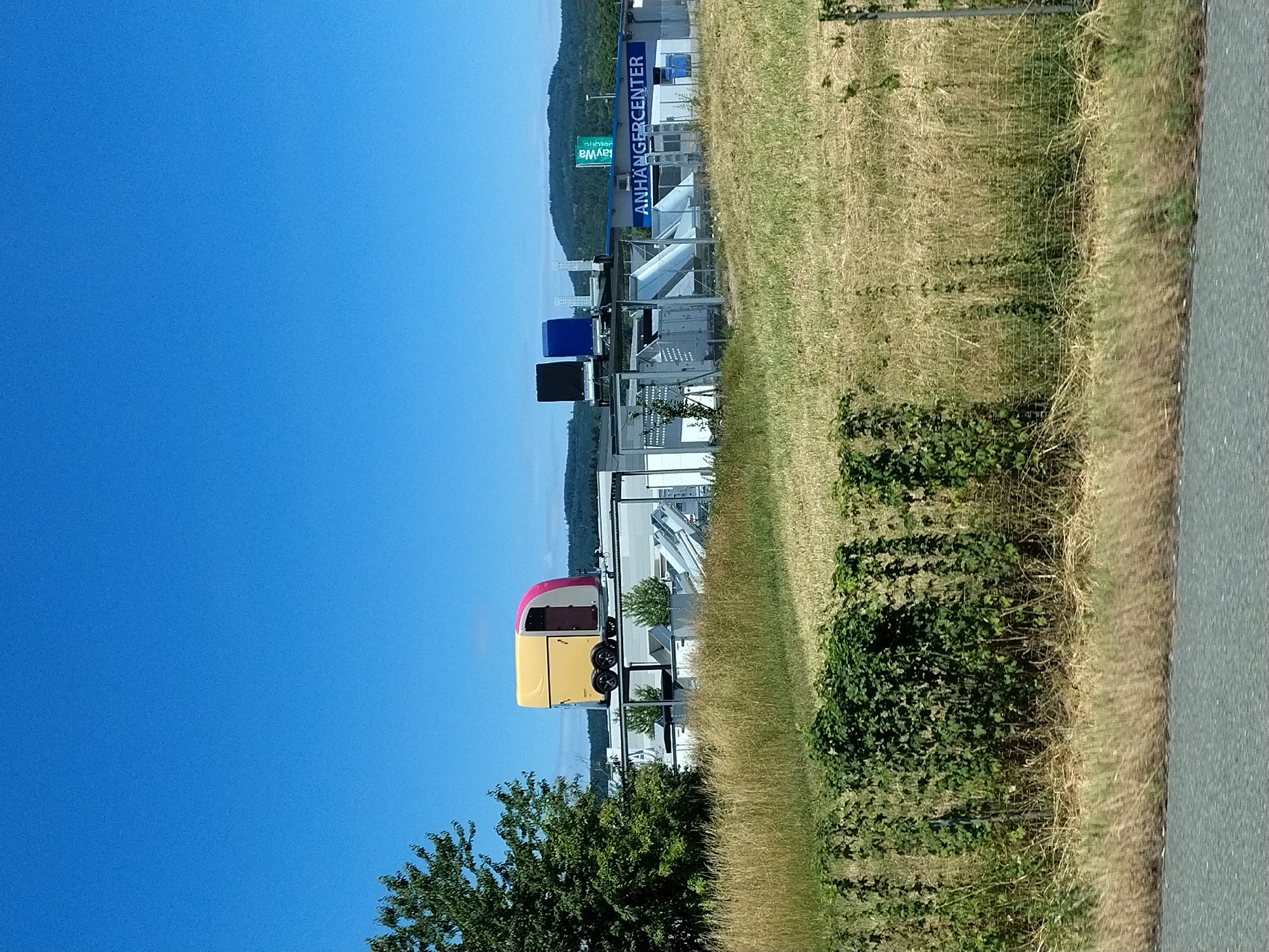 In Wörth an der Donau, Germany, a yellow and pink trailer, resembling a horse trailer, is prominently displayed on the roof of a building. To the right of the trailer, two dark blue and black rectangular structures are also situated on the building's roof. The building itself is a low-rise commercial structure with a white facade and a blue trim, featuring a sign that reads "ANHÄNGERCENTER". Adjacent to the building, a green sign with white lettering that says "StayWa" is visible. The scene is set against a backdrop of a clear blue sky and rolling green hills in the distance. In the foreground, a field of dry, golden grass stretches out, bordered by a dense green hedge. The image appears to be taken from a roadside perspective.
