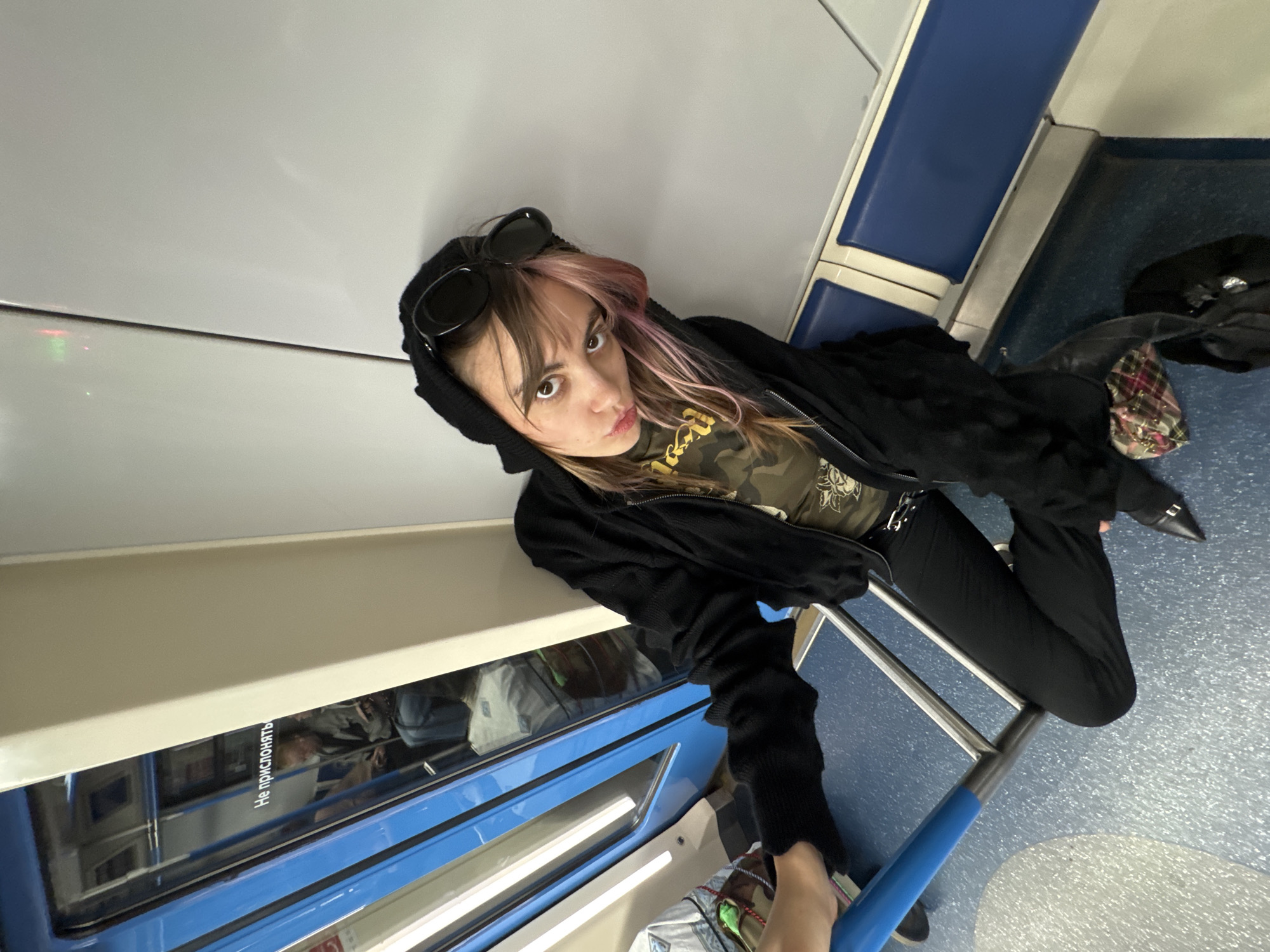 A young woman with pink-streaked hair and dark clothing is seated in a Moscow Metro train car. She wears a black hooded jacket, sunglasses perched on her head, a camouflage t-shirt, black pants, and pointed black boots. Her left hand rests on a blue pole and a metal railing. The train car's interior is visible, with blue seats and light-colored walls. A reflection in the window shows another passenger.