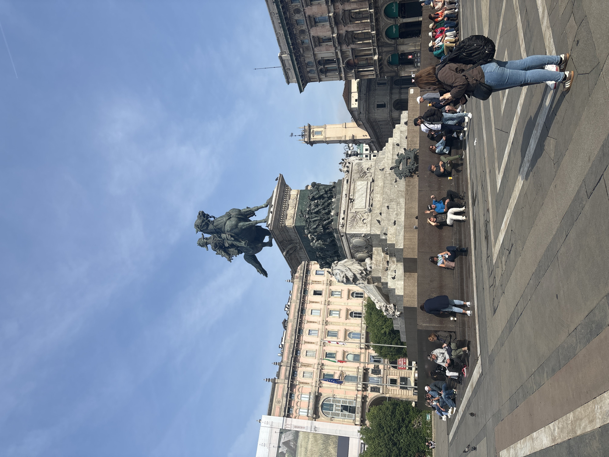 The image shows a street scene in Milan, Italy, featuring the equestrian statue of Victor Emmanuel II. The statue is situated in a plaza, with people sitting on the steps surrounding the monument. In the background are buildings, including those with arcades and a tower. The sky is clear and blue. People are sitting and standing around the statue.