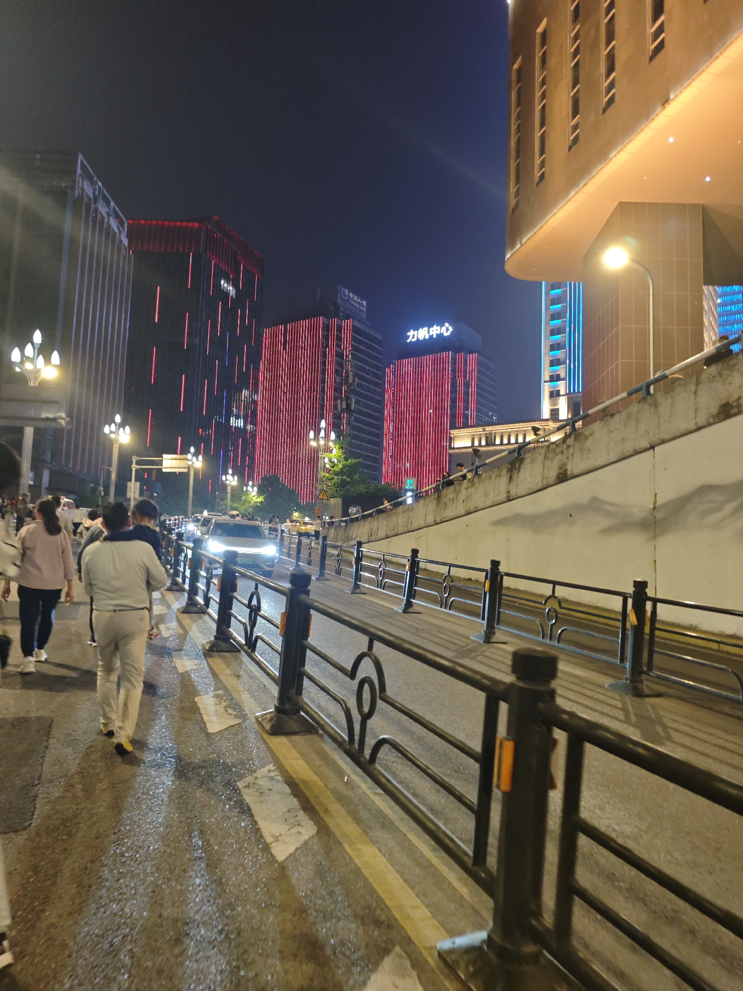 A nighttime street scene in 江北城街道, China, shows people walking along a sidewalk next to a street with cars. Buildings illuminated with red and blue lights are visible in the background. A black metal railing separates the sidewalk from the street. Streetlights illuminate the area, and the ground appears wet.