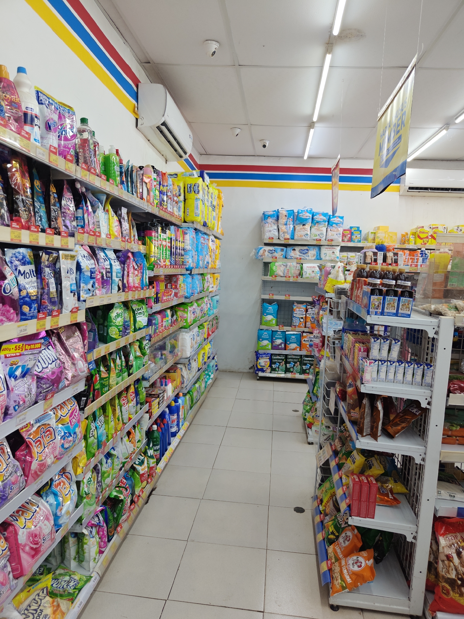 The image depicts the interior of a well-lit convenience store, likely a minimarket, with aisles stocked with various consumer goods. The store features white walls with red, blue, and yellow stripes near the ceiling, and a tiled floor. Fluorescent lights illuminate the scene from above, and an air conditioning unit is visible mounted on the wall.
The shelves are organized with products such as laundry detergent, fabric softener, baby diapers, food items like snacks and beverages, and cleaning supplies. Products are predominantly packaged in colorful bags and boxes. Some shelves have price tags.
On the left side of the image, shelves are filled with laundry detergents and fabric softeners, some with prominent branding like "Molto." Further down the aisle, more cleaning products and personal care items are visible.
In the center and right of the image, a separate shelving unit displays stacks of baby diapers on the upper shelves, followed by beverages and other packaged goods. The lower shelves of this unit and an adjacent display rack on the right are filled with various snack items, including bagged chips and cookies.
The scene is devoid of any people. The overall impression is one of a typical retail environment stocked for everyday consumer needs. Based on the prominent signage and product brands, the location is likely in Indonesia. Given the provided context, this store is situated in Klampokan, Indonesia.