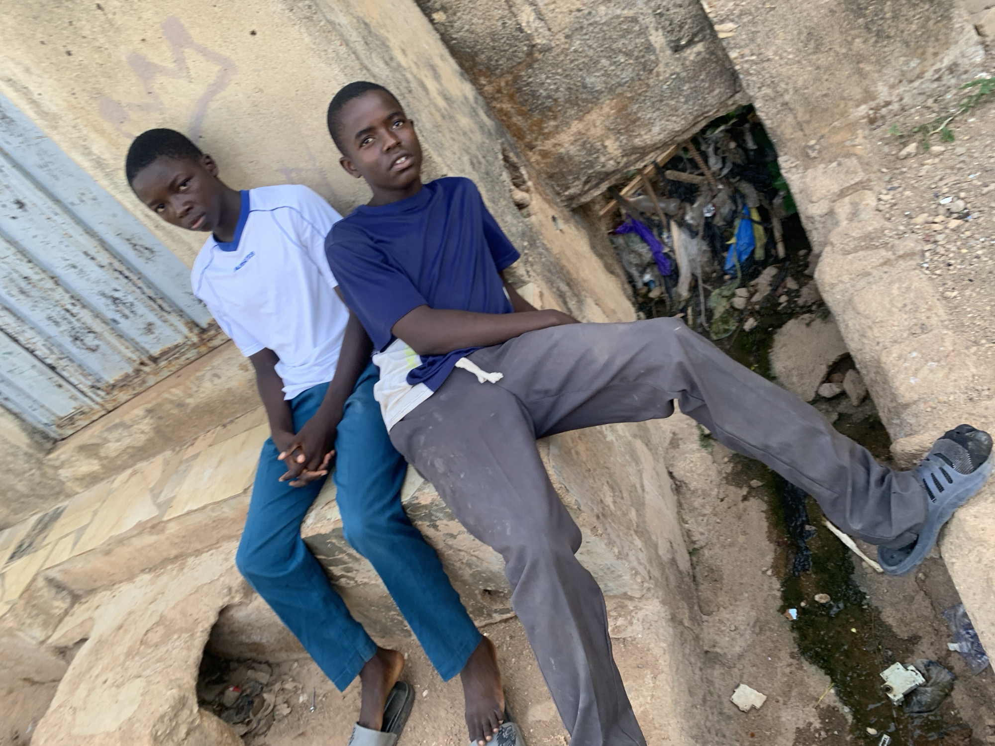 Two teenage boys are sitting on a concrete structure, likely a drainage ditch or culvert, in an urban environment. The boy on the left wears a white t-shirt with blue trim and blue jeans. He is barefoot, wearing gray sandals. The boy on the right wears a dark blue t-shirt and gray pants. He wears a dark colored Adidas sneaker on his right foot and a sandal on his left foot. They are both looking towards the camera. Debris and trash are visible in the drainage ditch. The location is possibly in Jos, Nigeria, based on clothing and the environment.