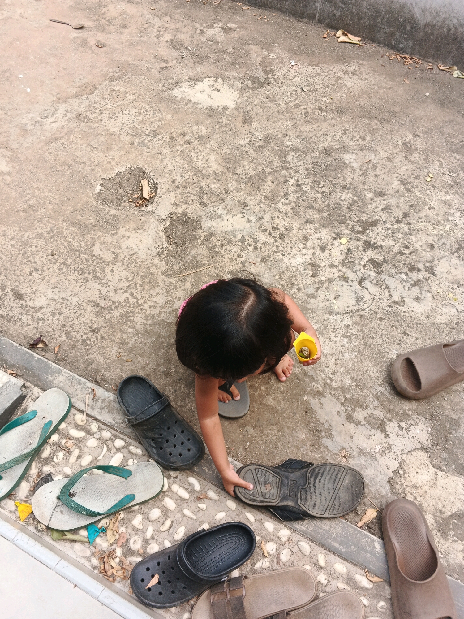 A high-angle, outdoor shot shows a young child, with dark hair and wearing a pink shirt, interacting with the ground. The child is holding a small yellow cup in their right hand, which contains some small items. The child's left hand is reaching towards a black sandal on the ground. The ground is a rough, concrete surface with scattered debris, including leaves and pebbles. Several pairs of footwear are visible around the child, including flip-flops, sandals, and clogs. The scene appears to be in a residential or outdoor play area in Kadu, Indonesia.