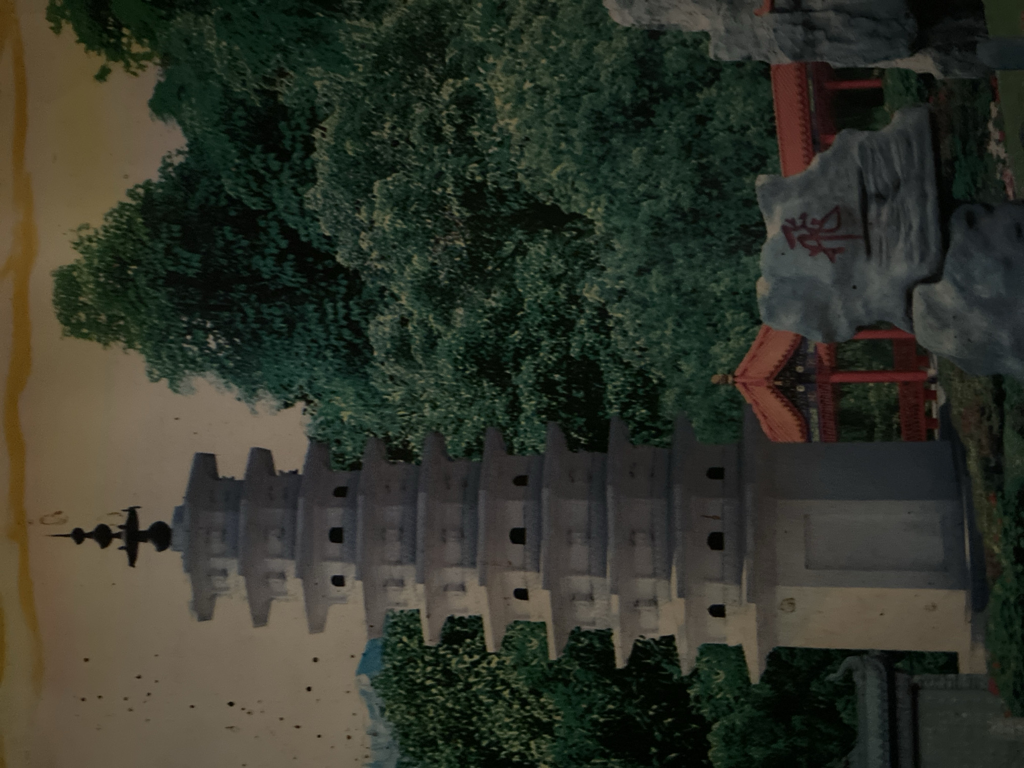The image depicts a picturesque scene featuring a multi-tiered pagoda in a lush, green environment. The pagoda, predominantly white, has several levels, each with projecting eaves and small, dark openings. It is situated in what appears to be a botanical garden or park. In the background, dense foliage and trees create a natural backdrop. To the right of the pagoda, a traditional Asian-style building with a red roof and intricate details is visible. In the foreground, a large, grey rock formation stands out, with a red Chinese character, likely "飛" (fēi), meaning "fly," painted on its surface. This character is positioned centrally on the rock. The overall scene suggests a serene and culturally inspired setting. This specific pagoda and its surrounding landscape are part of the Japanese Tunnel in Davao City, Philippines, a historical site that was once a Japanese military headquarters during World War II. The area has since been developed into a park and tourist attraction, incorporating elements of Japanese and other Asian architectural styles, including pagodas, as seen in this image.