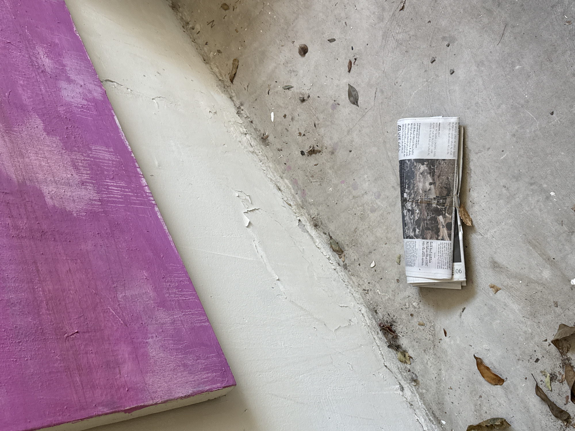 An indoor, ground-level view focuses on a folded newspaper lying on a concrete floor. The newspaper is oriented horizontally and is positioned towards the right side of the frame. The concrete floor is dusty and appears somewhat worn, with scattered debris, including dry leaves and small pieces of plaster. To the left and top of the frame, a wall painted in a textured, uneven, vibrant pink color angles diagonally downwards. Below the pink surface, the wall transitions to a rough, off-white textured finish that meets the floor at a distinct junction. The scene suggests an unfinished or neglected interior space, possibly a studio or a room undergoing renovation. The newspaper contains visible text and images, with some of the text appearing to be in Hebrew script. The overall lighting is neutral, with no strong shadows, indicating diffuse illumination. The image was taken in Tel-Aviv, Israel.