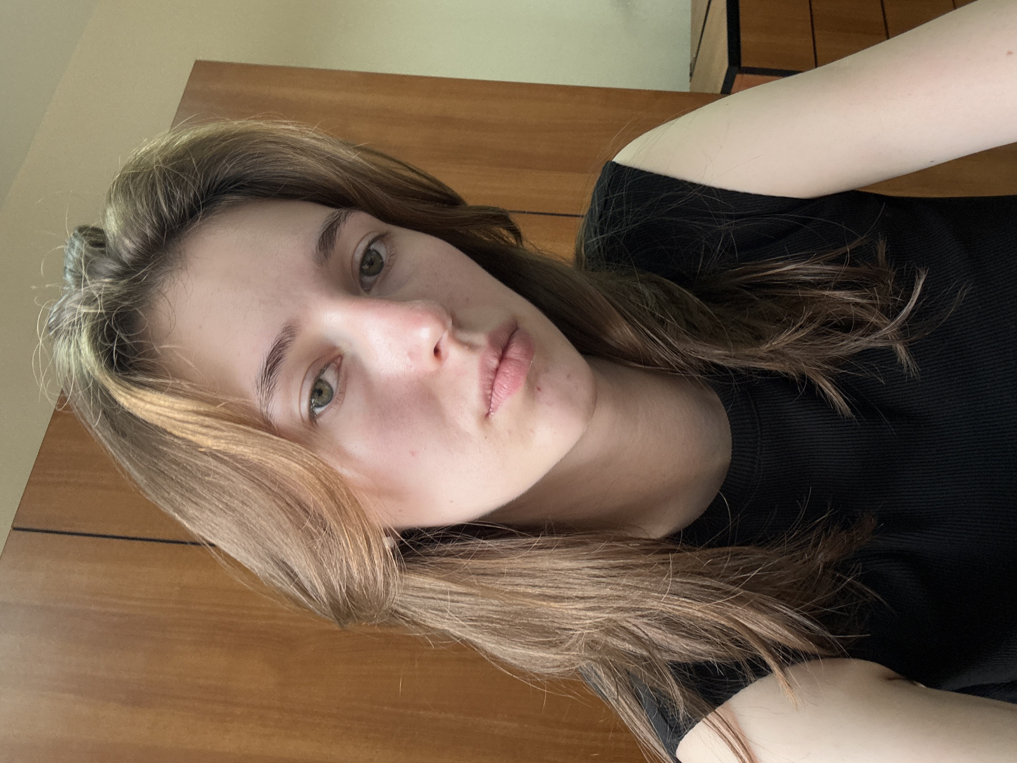 The image shows a close-up, waist-up, selfie-style portrait of a young woman with long, light brown hair, fair skin, and green eyes. She is wearing a black, sleeveless, ribbed top. Her hair is parted down the middle and falls loosely around her face and shoulders. Her skin appears to have some minor blemishes. The background consists of a wooden wardrobe and a light-colored wall. The lighting suggests an indoor setting, possibly a bedroom or living area. The image composition focuses on the subject's face and upper body. No specific details of the city of Perm, Russia, are visible in the image.
