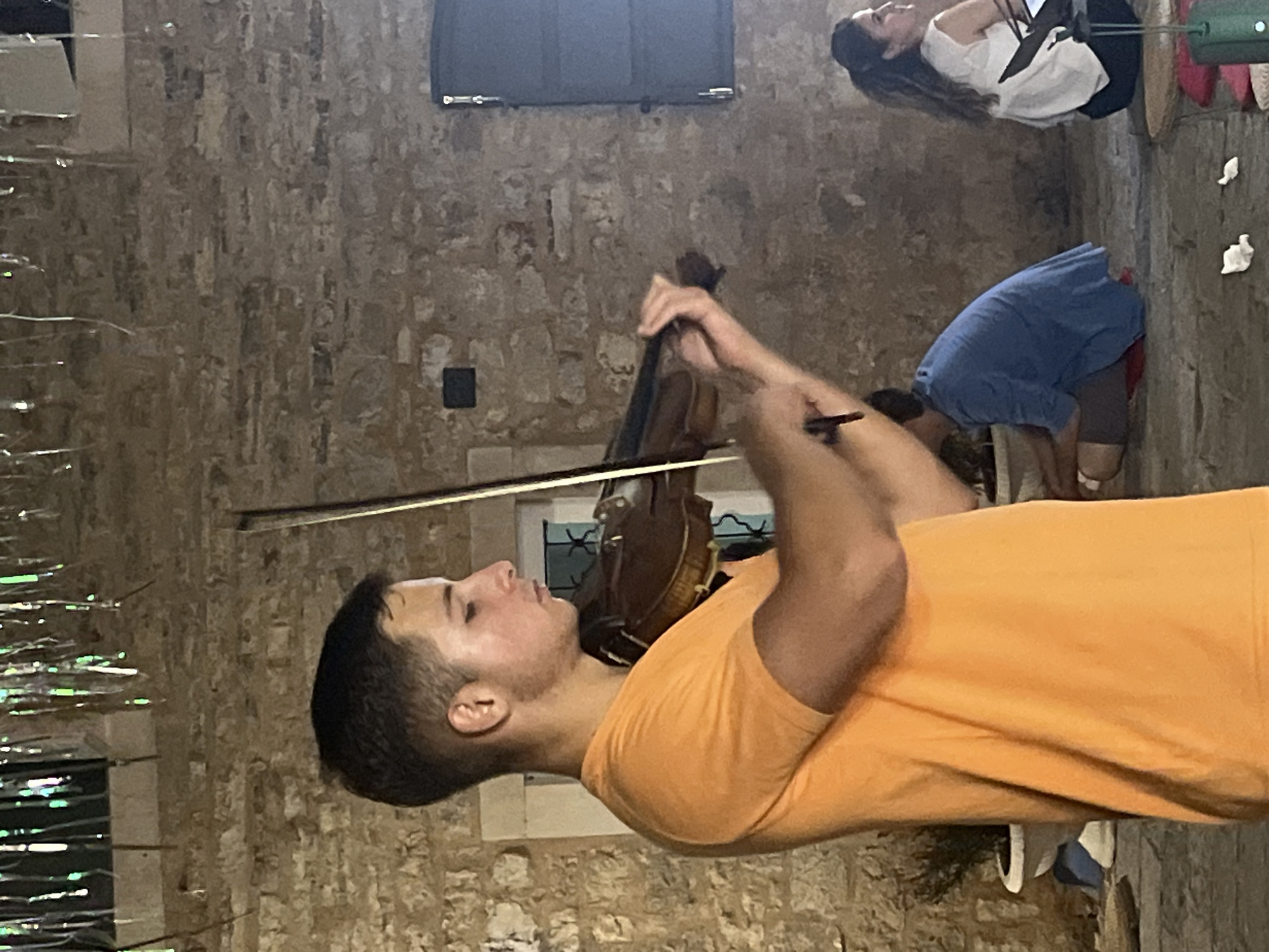 A young man in an orange t-shirt plays the violin in front of a rough stone wall. His face is in profile, and his eyes are closed as he concentrates on his music. His left hand is on the neck of the violin, and his right hand is on the bow. A young person in a blue shirt and shorts is sitting on the ground behind him, and a woman in a white top and dark skirt is seated to the right, looking away from the camera. The scene appears to be outdoors, possibly in a courtyard, with decorative silver streamers hanging from above. The setting is the old town of Budva, Montenegro, near the Church of St. John.