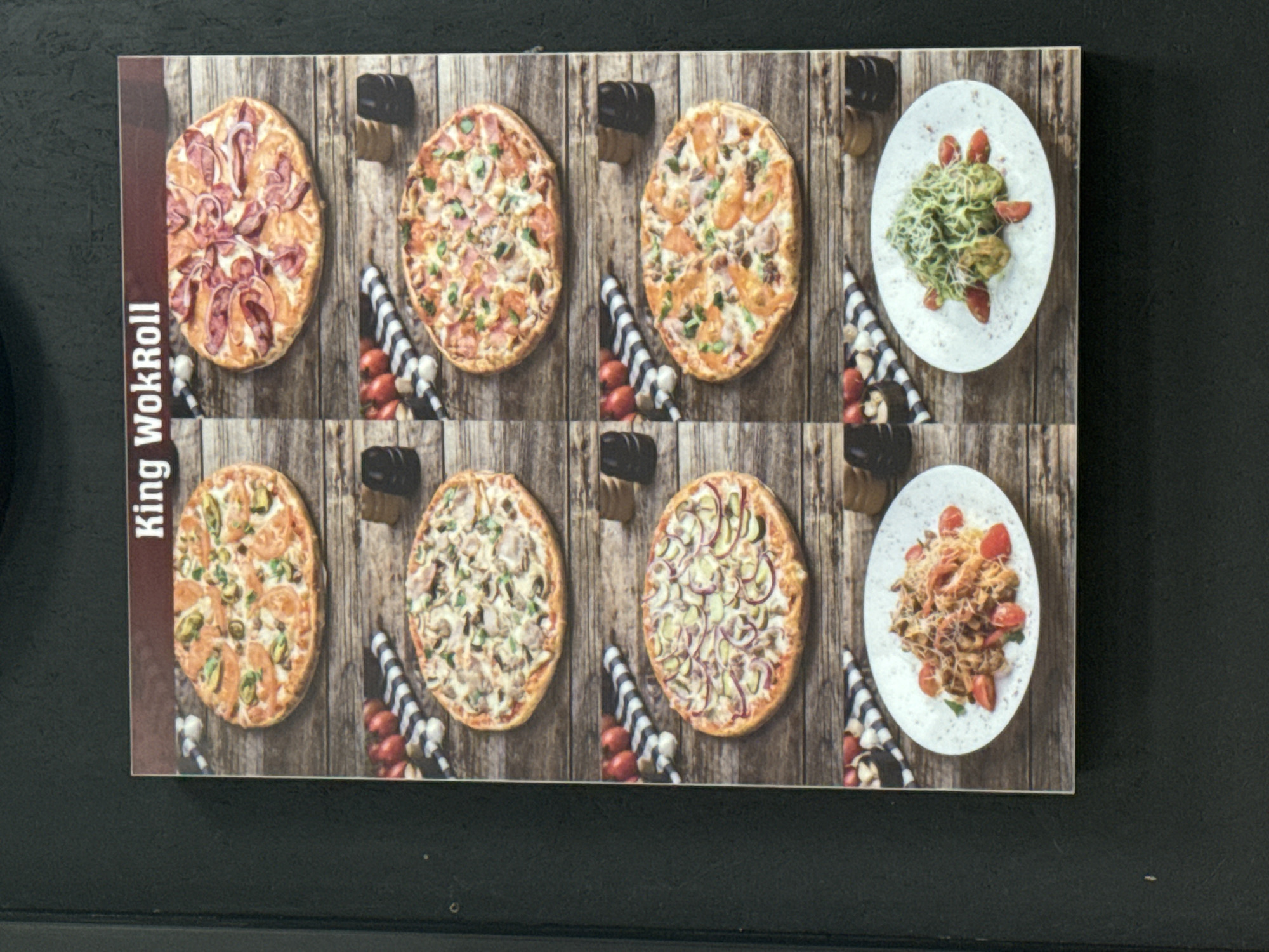 The image shows a printed menu for "King WokRoll", likely a restaurant or delivery service, displayed against a black surface. The menu features images of several dishes including pizzas with various toppings (e.g., pepperoni, mushrooms, vegetables), and pasta dishes with tomatoes and cheese. The image is set against a rustic wooden plank background. No people are visible. The location is Mytishchi, Russia, based on the presumed origin of the image.