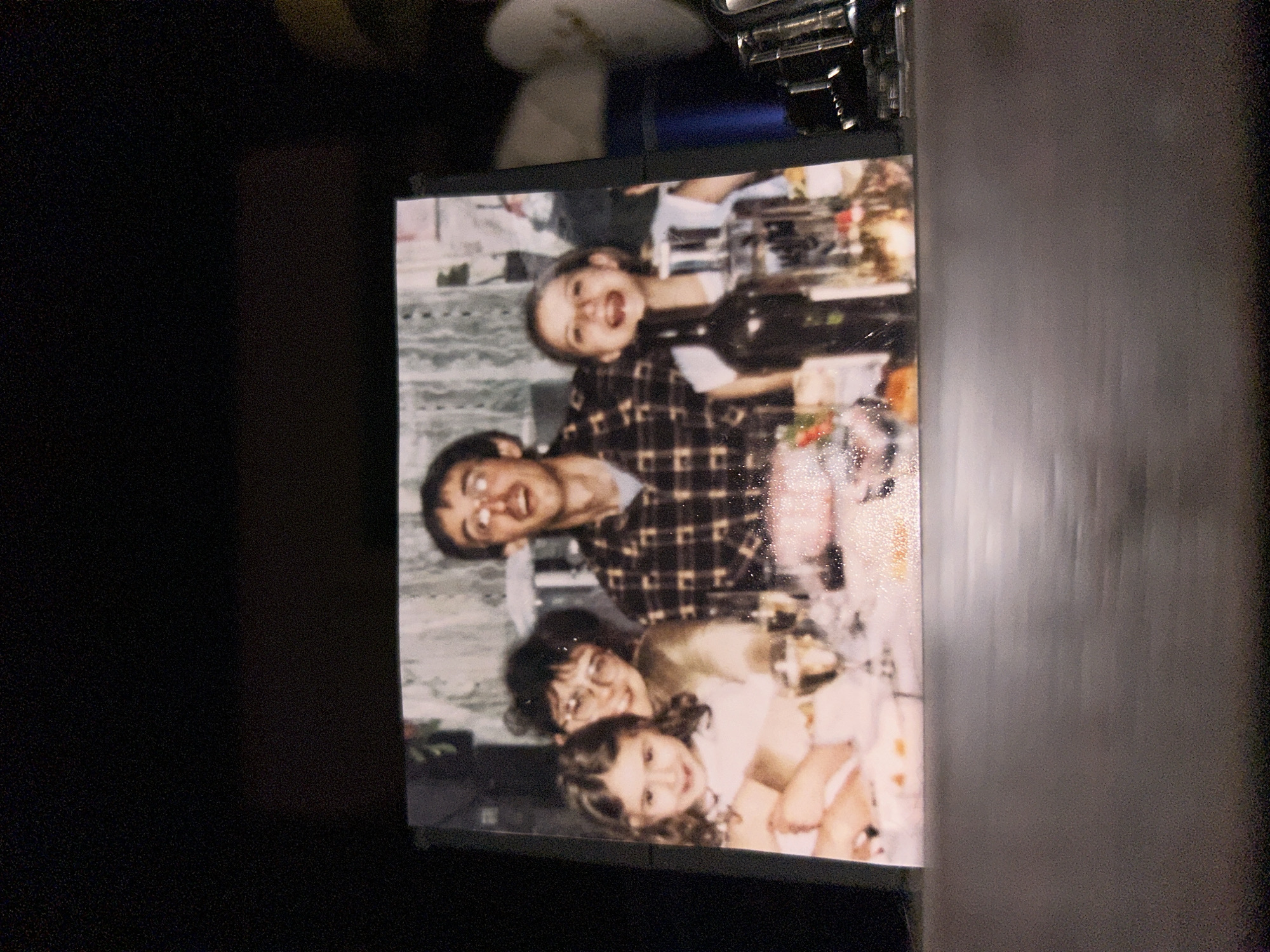 A photograph depicts a family of four, with an adult man in a brown plaid shirt, an adult woman, and two younger females. They are positioned around a table with food and a wine bottle. The exact location is unknown.